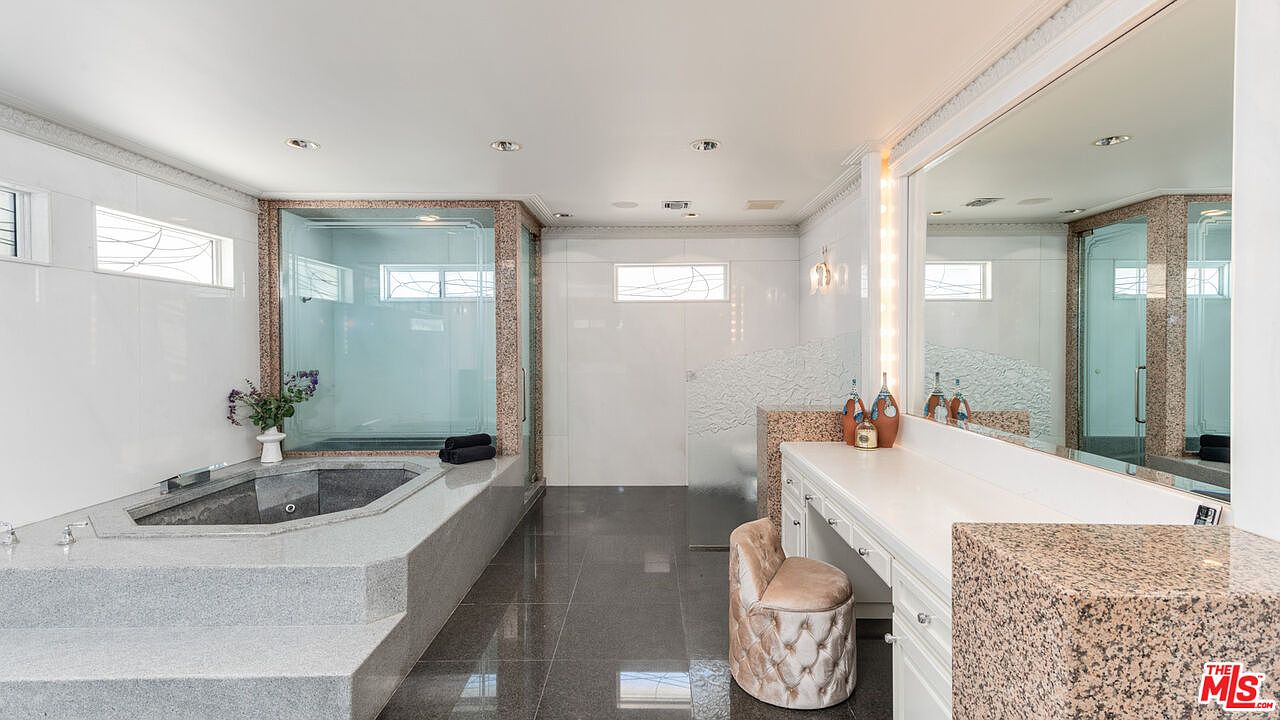This is a luxurious primary bathroom featuring a large, elevated jacuzzi tub with gray granite steps and a glass-enclosed shower. The vanity area includes a makeup station with a tufted stool and a large mirror with vanity lighting. The flooring is dark gray tile, and the overall design is elegant and spacious.