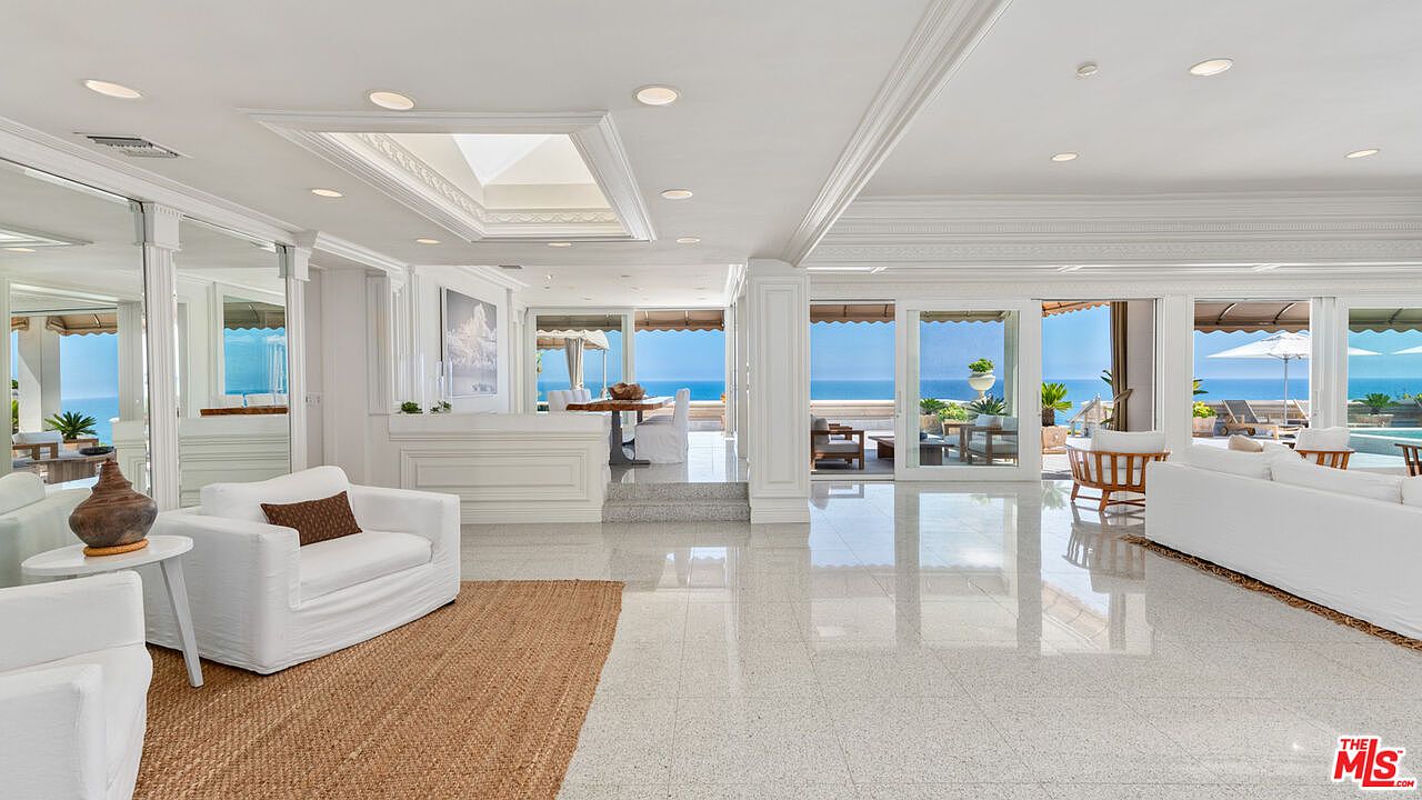 This is a luxurious living room with a stunning ocean view. The room features white furniture, a jute rug, and large sliding glass doors that open to a patio or deck. The interior design is elegant and bright, creating a serene and inviting atmosphere.