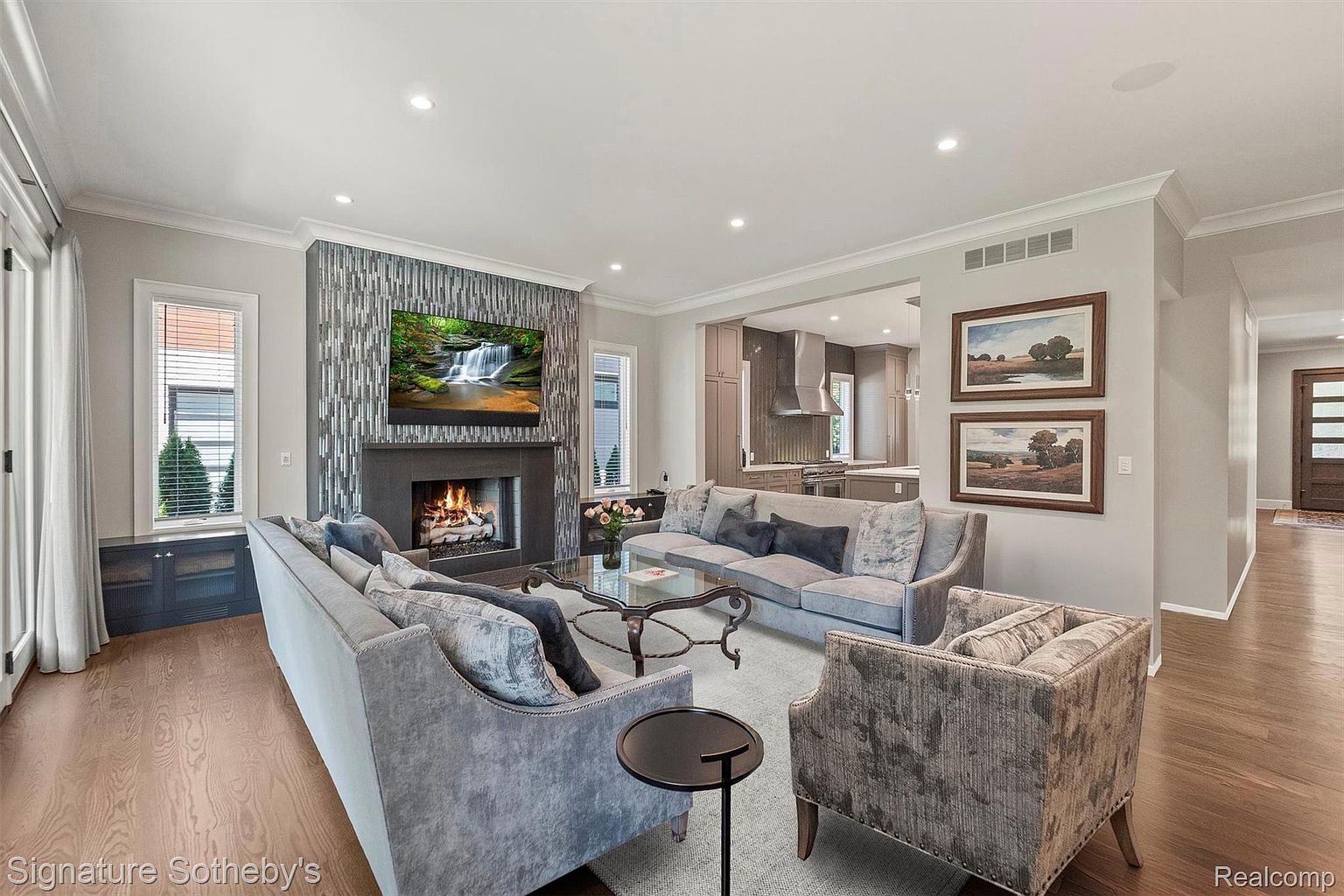 This is an interior shot of a living room featuring two sofas, an armchair, a fireplace with a television mounted above it, and two framed paintings on the wall. The room is decorated in neutral tones with gray and silver accents, creating a sophisticated and comfortable atmosphere. The hardwood floors and area rug add warmth and texture to the space.