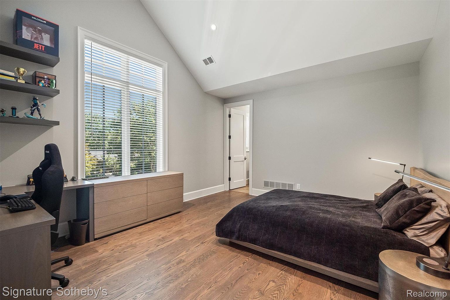 This is a well-lit bedroom featuring hardwood floors and light gray walls. A large window with blinds provides natural light, complemented by a modern desk area with shelves. The bed is neatly made with dark bedding, and a side table adds a touch of elegance, creating a serene and inviting space.