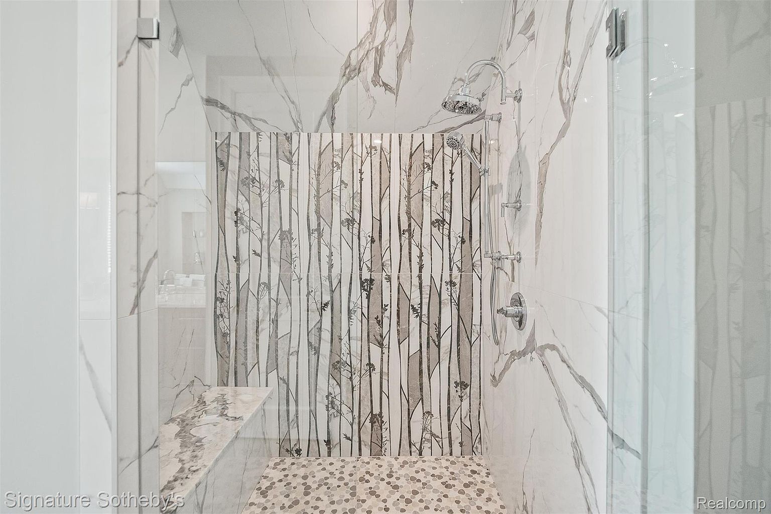 This is a luxurious primary bathroom featuring a walk-in shower with marble-like tile walls and a decorative tile accent wall. The shower includes a rainfall showerhead and a handheld sprayer, with a built-in bench for added comfort. The overall impression is modern and spa-like, emphasizing high-end finishes and a serene atmosphere.