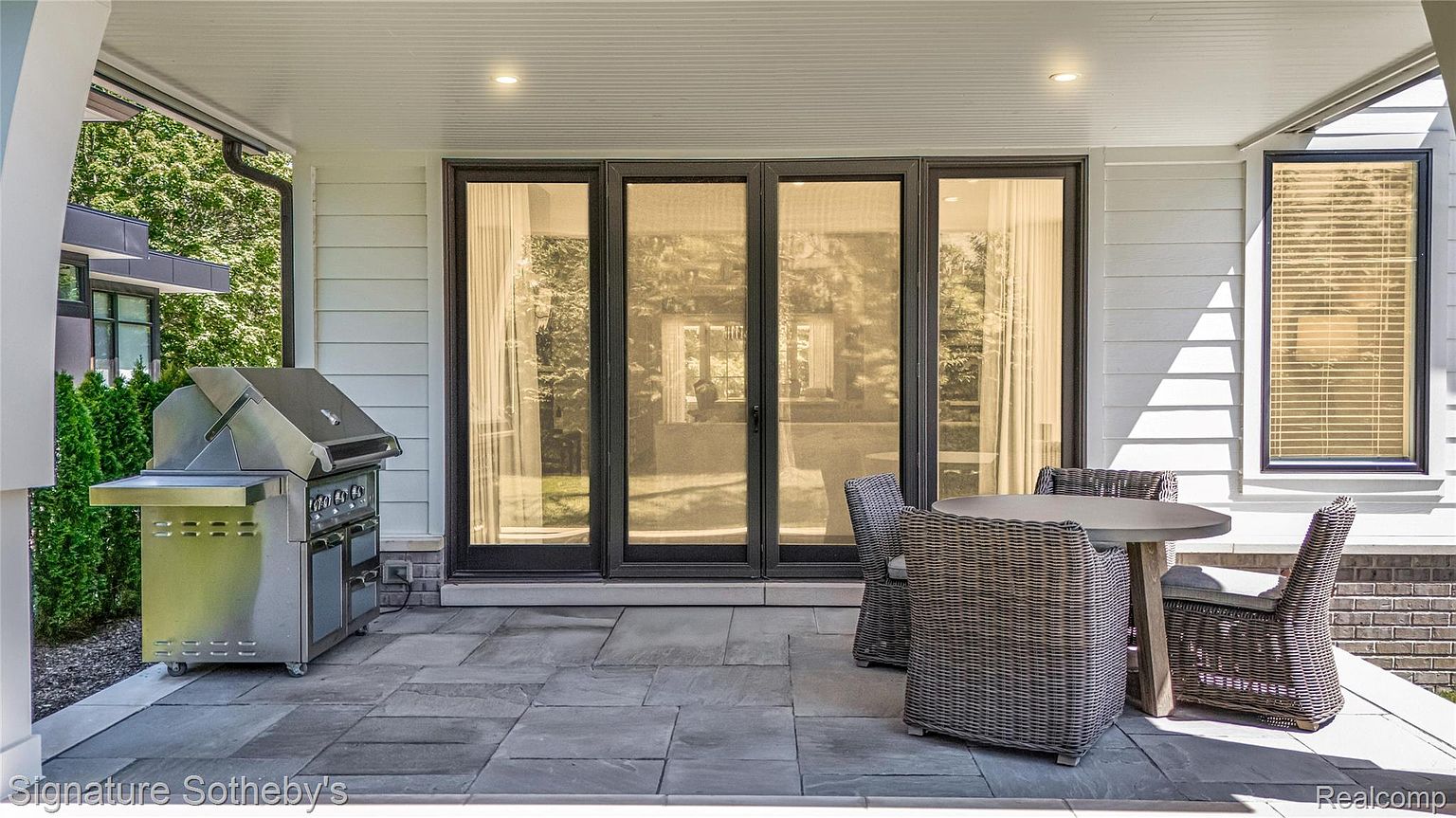 This image showcases a stylish outdoor patio area, featuring a built-in grill, a round table with wicker chairs, and a set of folding glass doors leading into the home. The patio is paved with large stone tiles, and the exterior of the house is clad in white siding. The scene suggests a comfortable and inviting space for outdoor dining and relaxation.