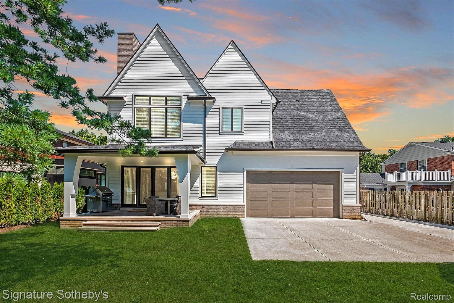 This is a front exterior view of a two-story house with white siding and a gray roof. The house features a covered porch with outdoor seating and a grill, a two-car garage with a brown door, and a well-maintained lawn. The overall impression is of a modern and well-kept family home.
