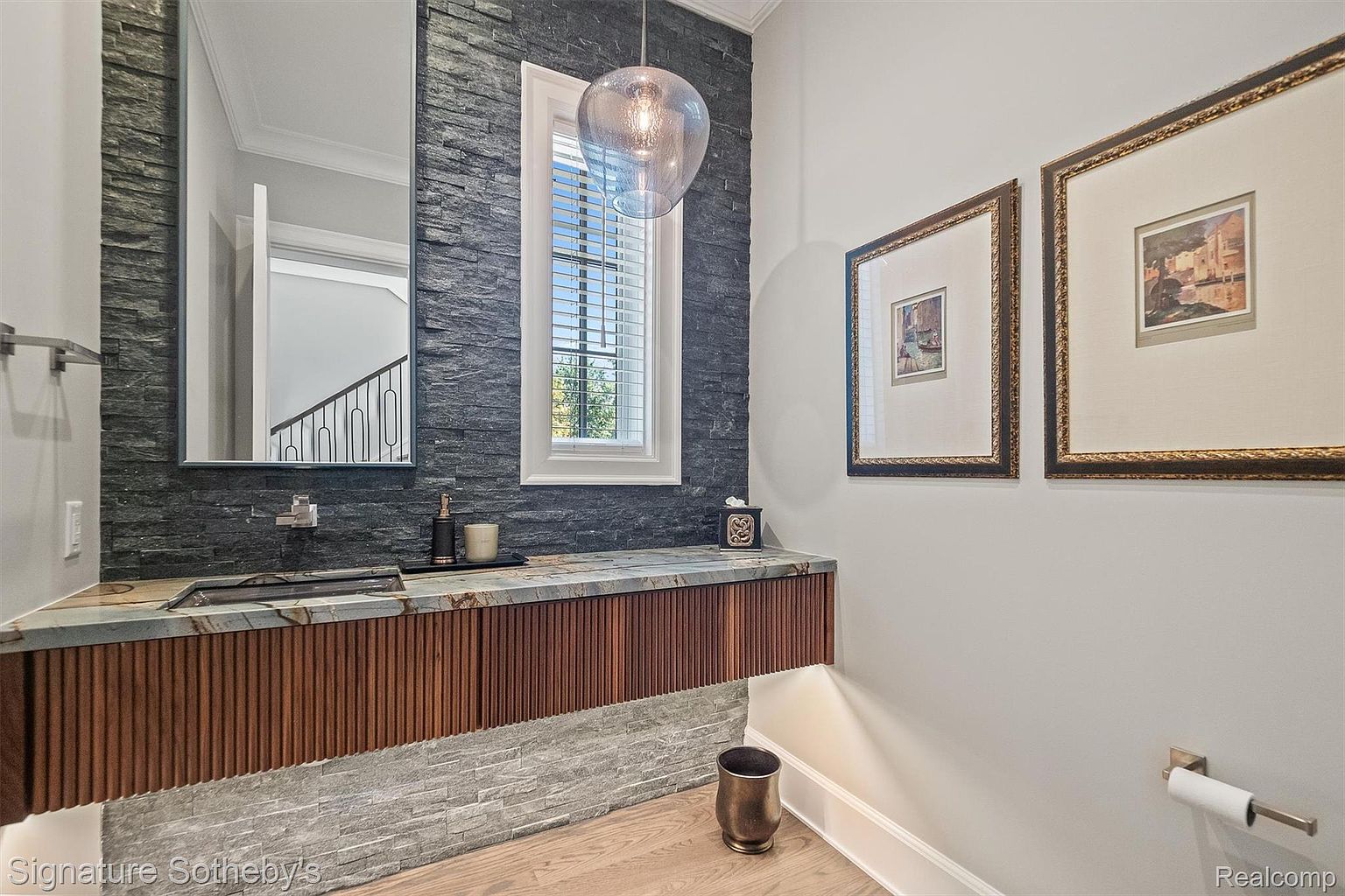 This is a stylish guest bathroom featuring a unique stone accent wall and a floating vanity with a stone countertop and wooden paneling. A modern mirror and pendant light add to the contemporary aesthetic. The room is well-lit and features neutral wall colors, complemented by framed artwork.