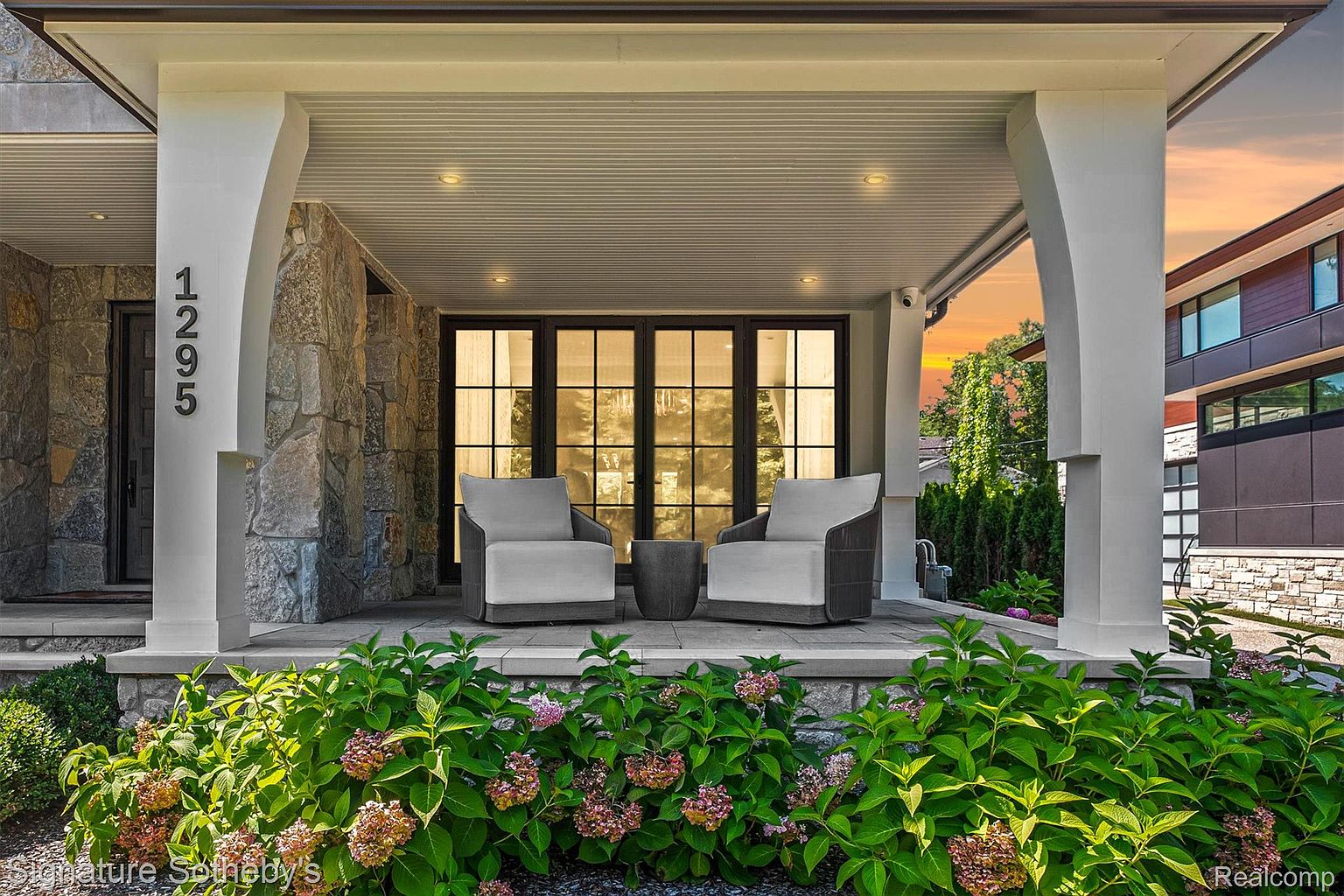 This image showcases a charming patio or balcony area of a home, featuring comfortable seating with two armchairs and a small table. The space is framed by lush greenery and stone accents, creating an inviting and relaxing outdoor living area. The architectural details, including the columns and stone facade, add to the property's curb appeal.