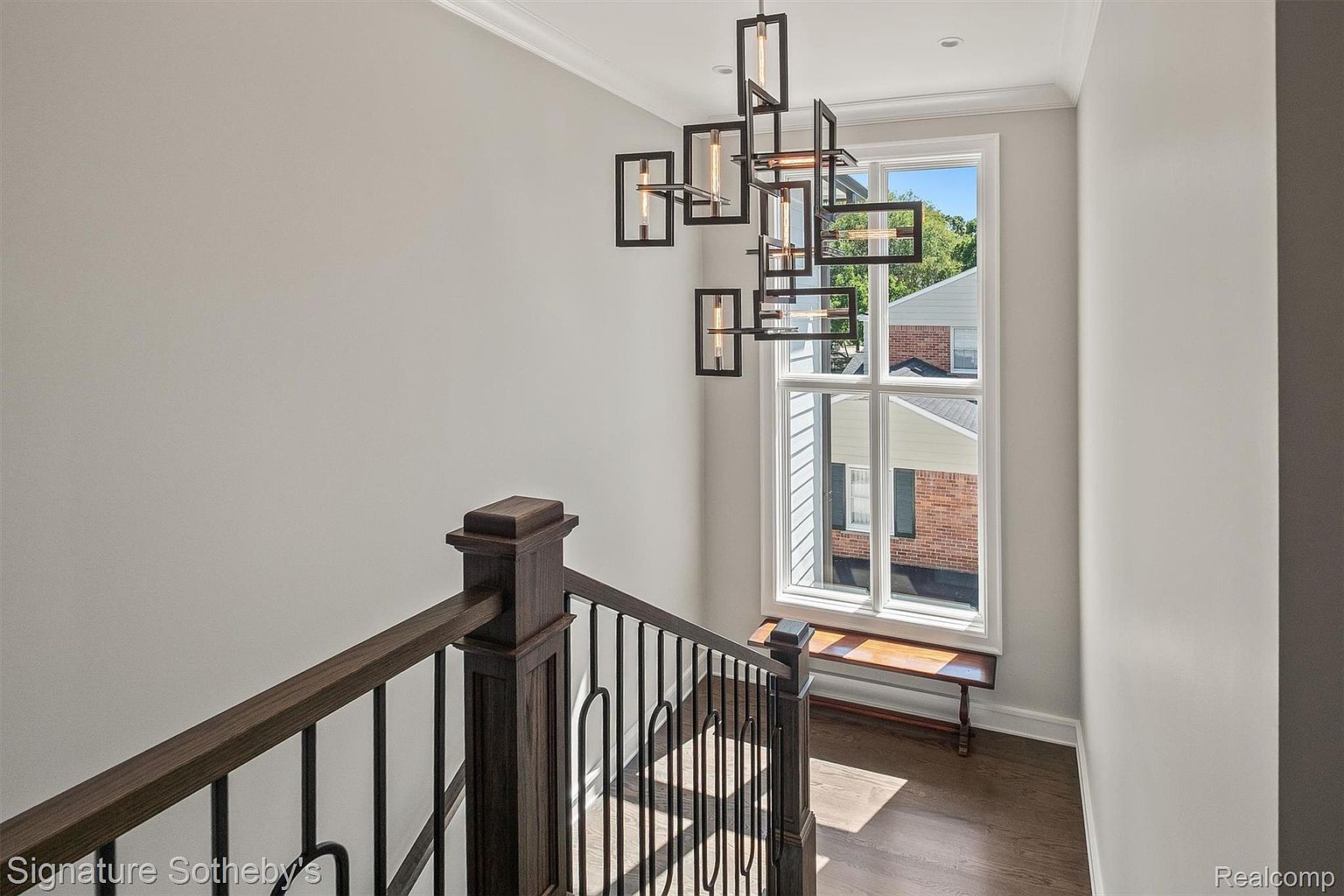 This image showcases a well-lit hallway or staircase landing featuring a modern chandelier and a large window that provides ample natural light. The dark wood railing and trim contrast with the light walls, creating a sophisticated and inviting atmosphere. A small bench sits beneath the window, offering a cozy seating area.