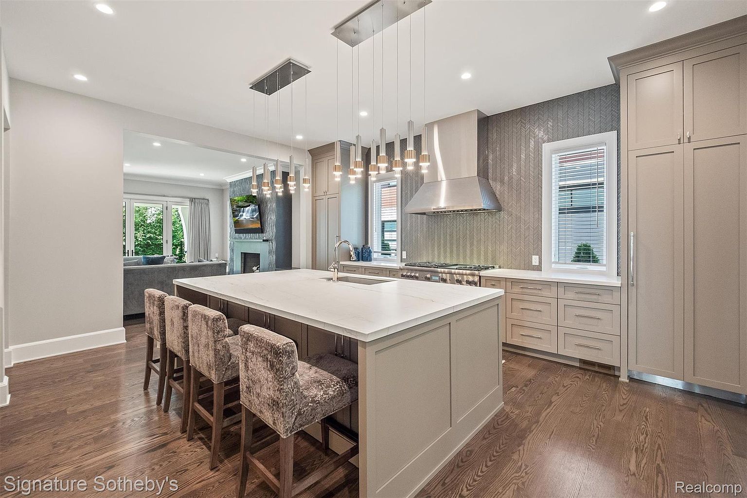 This is a well-lit, modern kitchen featuring a large island with seating, stainless steel appliances, and custom cabinetry. The kitchen has a neutral color palette with a textured backsplash and unique pendant lighting. The open layout connects to a living area, enhancing the sense of space and flow.