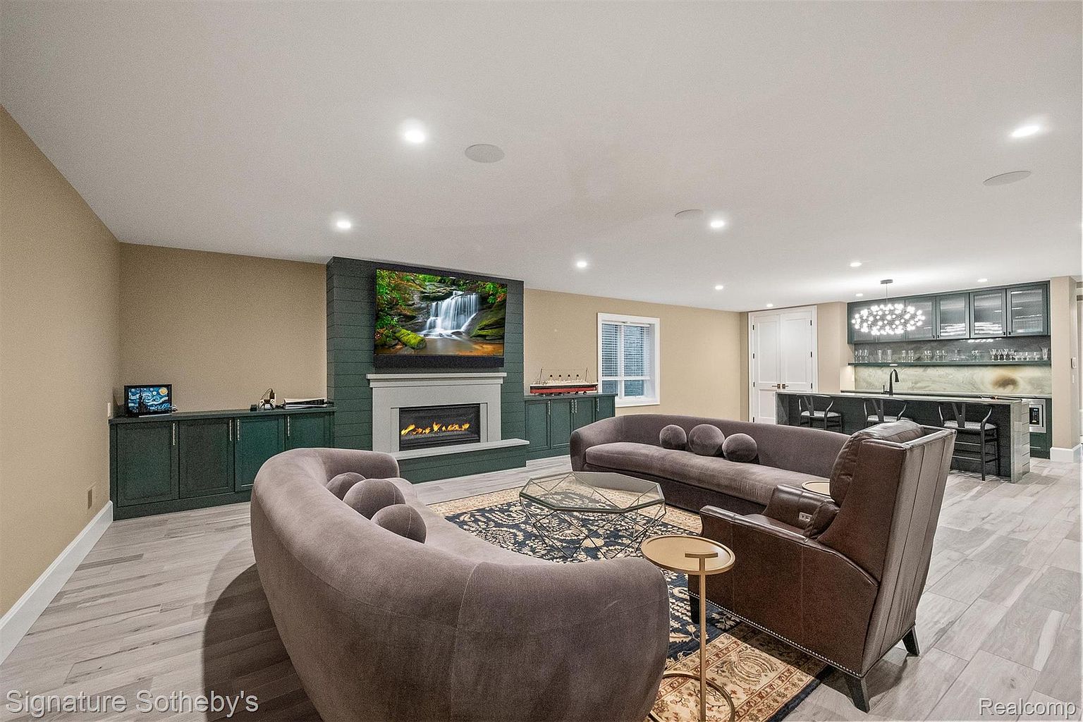 This is an interior shot of a living room featuring two curved sofas, a leather armchair, and a glass coffee table on a patterned rug. A fireplace with a mounted television is visible, along with built-in cabinetry and a bar area in the background. The room is well-lit with recessed lighting and has a modern, luxurious feel.
