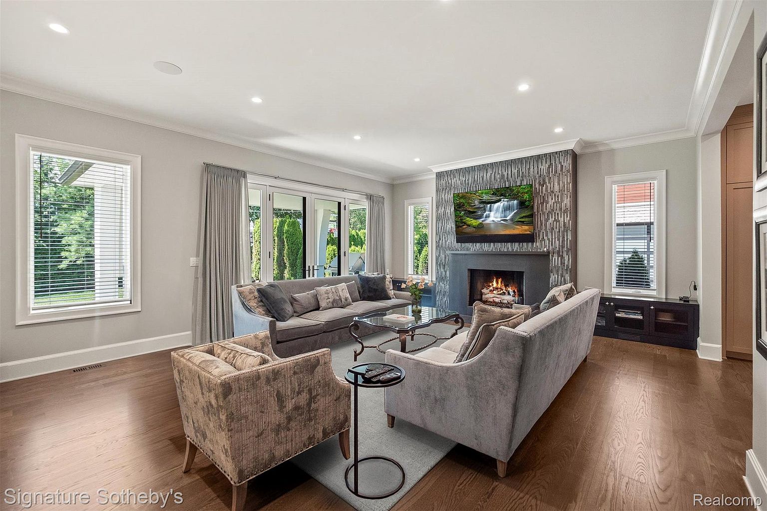 This is an interior shot of a living room featuring hardwood floors, a stone fireplace with a television mounted above, and two sofas facing each other. The room is well-lit with natural light coming through large windows and glass doors, complemented by recessed lighting. The overall impression is one of comfort and sophistication, ideal for relaxation and entertaining.
