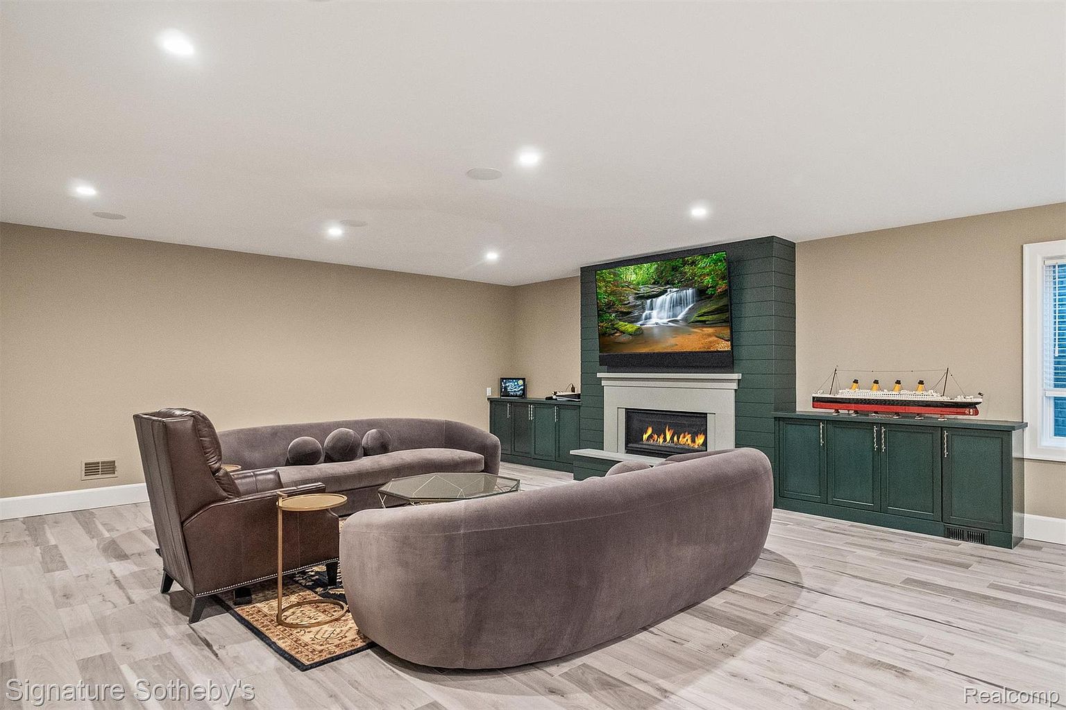 This is an interior shot of a living room featuring a modern design with neutral tones. The room includes two curved sofas, a leather armchair, a glass coffee table, and a fireplace with a large television mounted above. Built-in cabinetry flanks the fireplace, and the flooring appears to be light-colored wood-look planks, creating a cozy and stylish atmosphere.