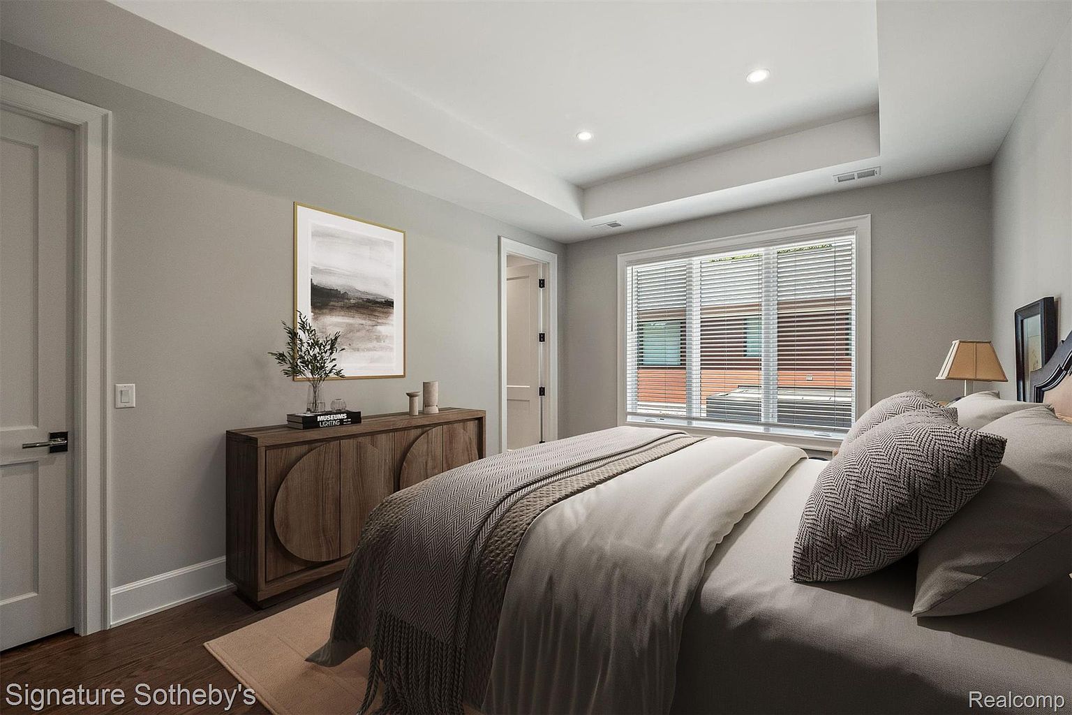 This is a primary bedroom featuring a neutral color palette with gray walls and hardwood floors. A large window with blinds provides natural light, and a wooden dresser with decorative items sits against one wall. The bed is neatly made with a gray comforter and decorative pillows, creating a serene and inviting atmosphere.