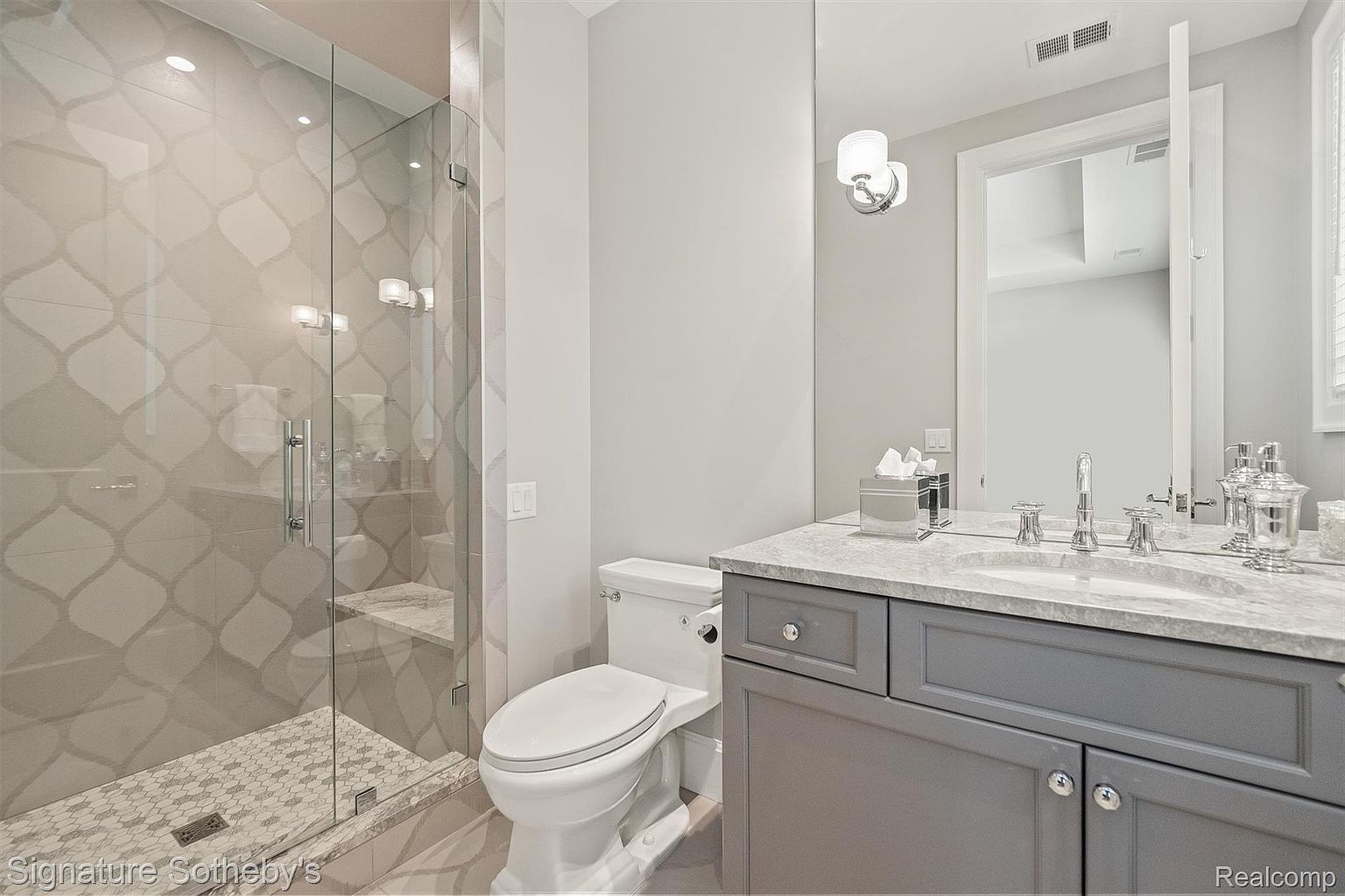 This is a well-lit bathroom featuring a glass-enclosed shower with patterned tile and a built-in bench. The vanity has a gray cabinet with a marble countertop, complemented by a large mirror and modern lighting fixtures. A white toilet is positioned next to the vanity, creating a functional and stylish space.