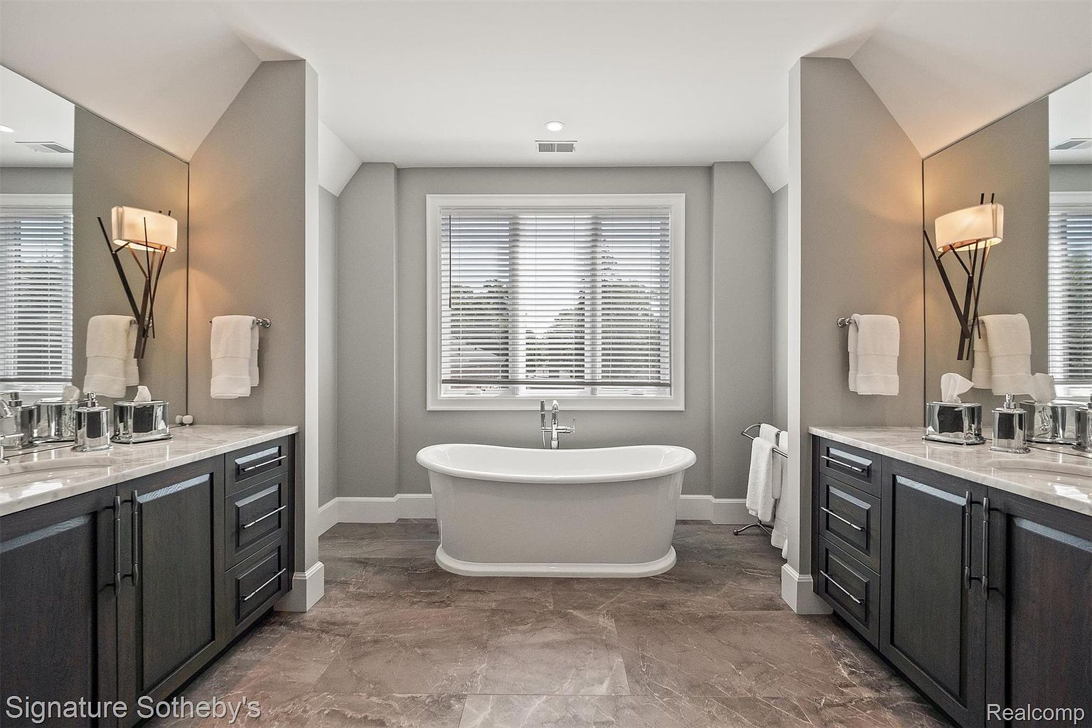 This is a luxurious primary bathroom featuring dual vanities with dark wood cabinetry and marble countertops. A freestanding bathtub sits beneath a window with blinds, providing natural light. The flooring is a polished stone tile, and the walls are painted in a neutral gray tone, creating a serene and elegant atmosphere.