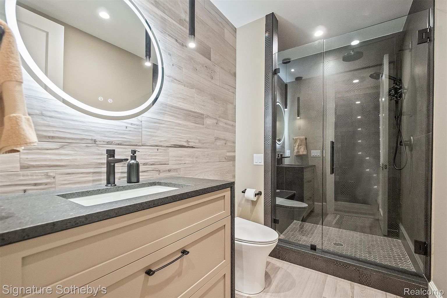 This is a modern primary bathroom featuring a vanity with a dark countertop and light-colored cabinetry, complemented by a round, illuminated mirror. A glass-enclosed shower with dark tiling and fixtures is visible in the background, along with a toilet. The overall design is sleek and contemporary, creating a luxurious and functional space.
