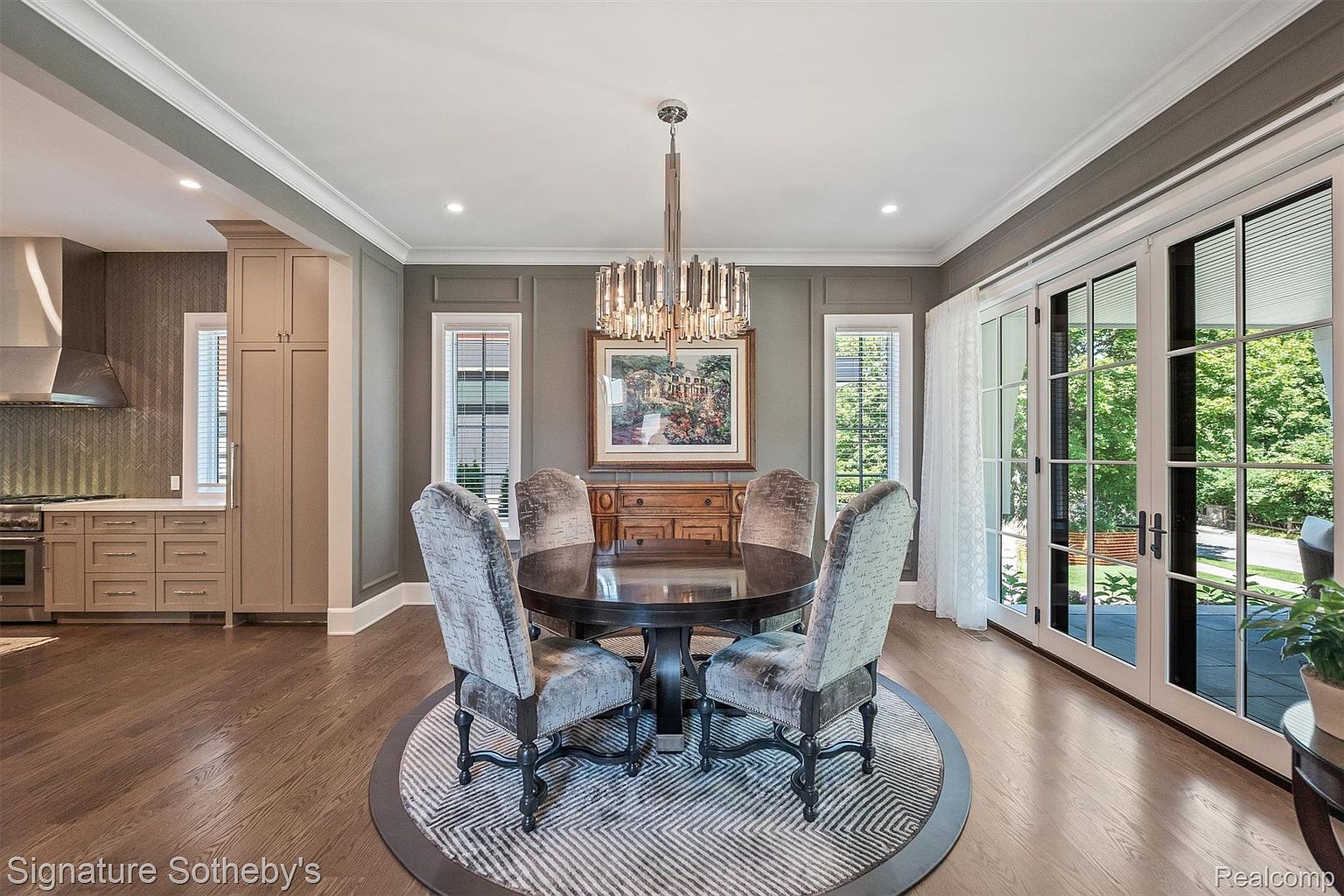 This is an interior shot of a dining room featuring a round wooden table with upholstered chairs set on a circular patterned rug. A modern chandelier hangs above the table, and a sideboard is positioned against the wall beneath a framed painting. Natural light floods the room through large glass doors, enhancing the elegant and inviting atmosphere.