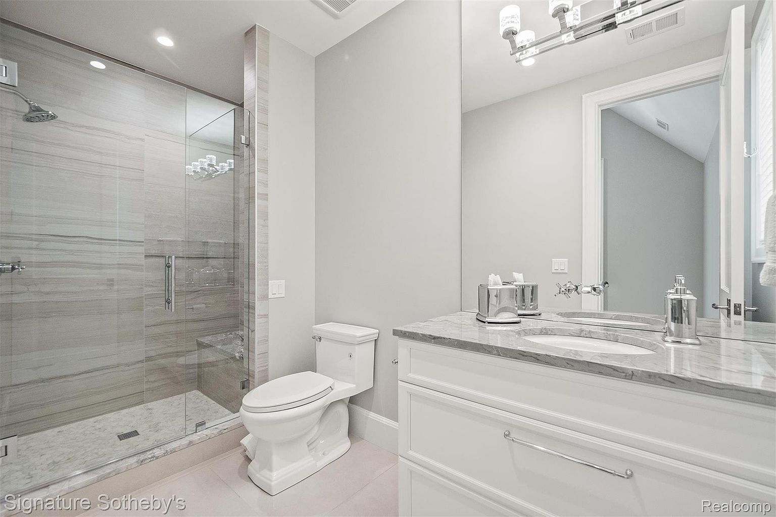 This is a well-lit bathroom featuring a glass-enclosed shower with gray marble-like tiling, a white toilet, and a white vanity with a marble countertop. A large mirror reflects the vanity area and a doorway leading to another room, enhancing the sense of space. The overall aesthetic is clean, modern, and luxurious.