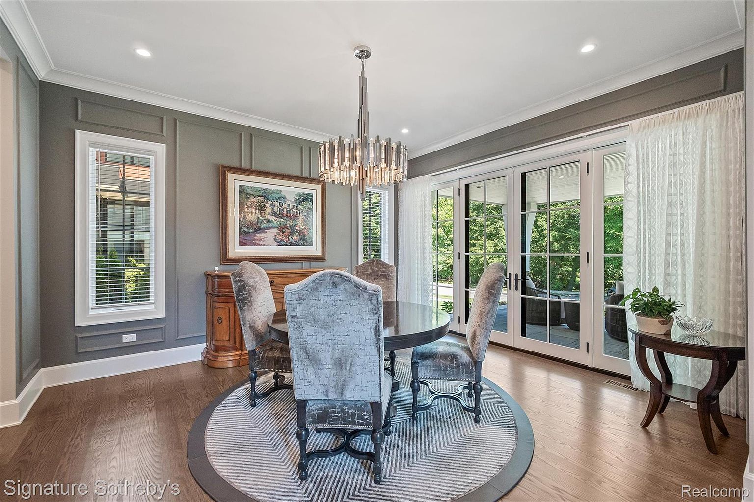 This is an interior shot of a dining room featuring a round dark wood table surrounded by four upholstered chairs with dark wood frames. A modern chandelier hangs above the table, and a painting adorns the wall above a wooden cabinet. The room has a sophisticated and elegant feel, enhanced by the gray walls, white trim, and hardwood floors.
