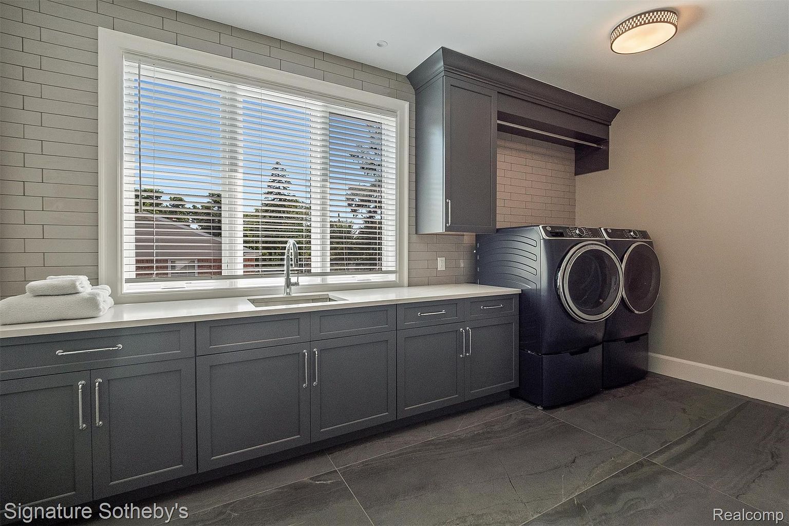 This is a well-appointed laundry room featuring gray cabinetry with sleek hardware, a white countertop, and a stainless steel sink beneath a large window with blinds. A modern, dark-colored washer and dryer set is positioned against the wall, complemented by matching upper cabinetry. The room is finished with gray tile flooring, creating a clean and contemporary space.