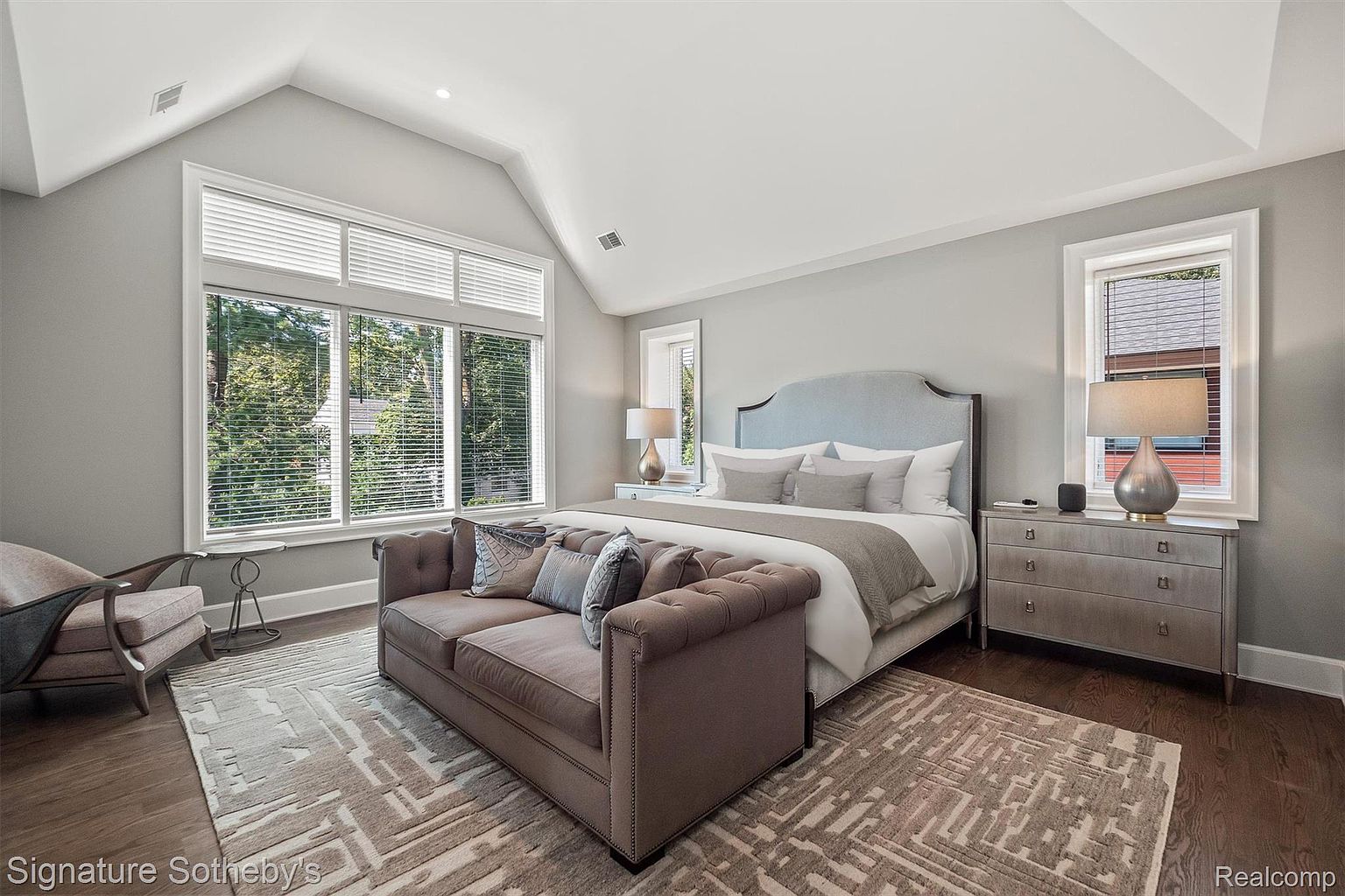 This is a primary bedroom featuring a large bed with a light blue headboard, several pillows, and a gray throw. A brown tufted sofa sits at the foot of the bed, and a dresser with lamps is positioned on the right side of the frame. Large windows provide natural light, and a patterned rug covers the hardwood floor, creating a cozy and inviting atmosphere.