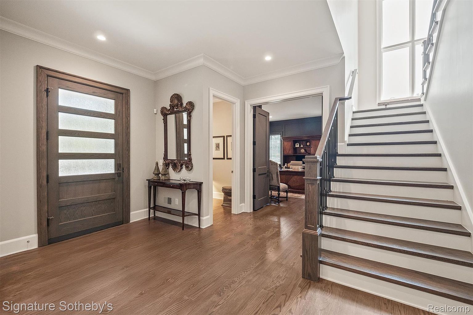 This is an interior shot of a home's entryway and staircase. The entryway features a wooden door with frosted glass panels, a decorative mirror above a small table, and hardwood flooring. The staircase has white risers and dark wood treads, leading to an upper level, creating a welcoming and elegant first impression.