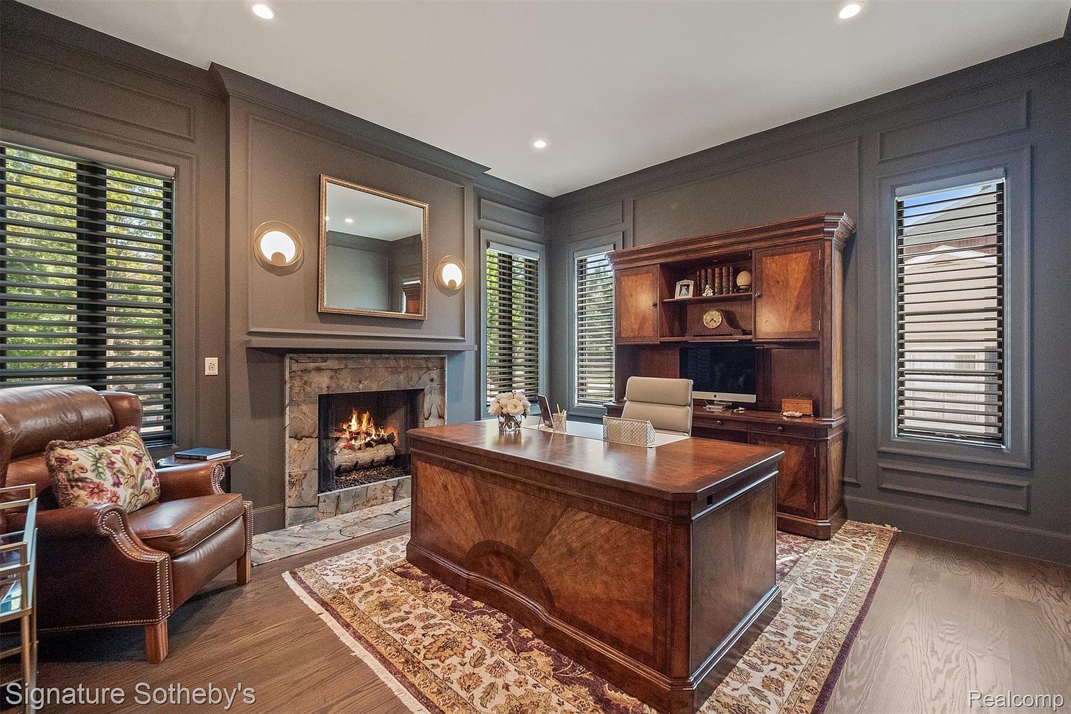 This is a well-appointed home office featuring a large wooden desk as the focal point, complemented by a built-in hutch and a comfortable leather armchair. A stone fireplace adds warmth and character to the room, while the dark walls and window blinds create a sophisticated and private atmosphere. The room is illuminated by recessed lighting and sconces.