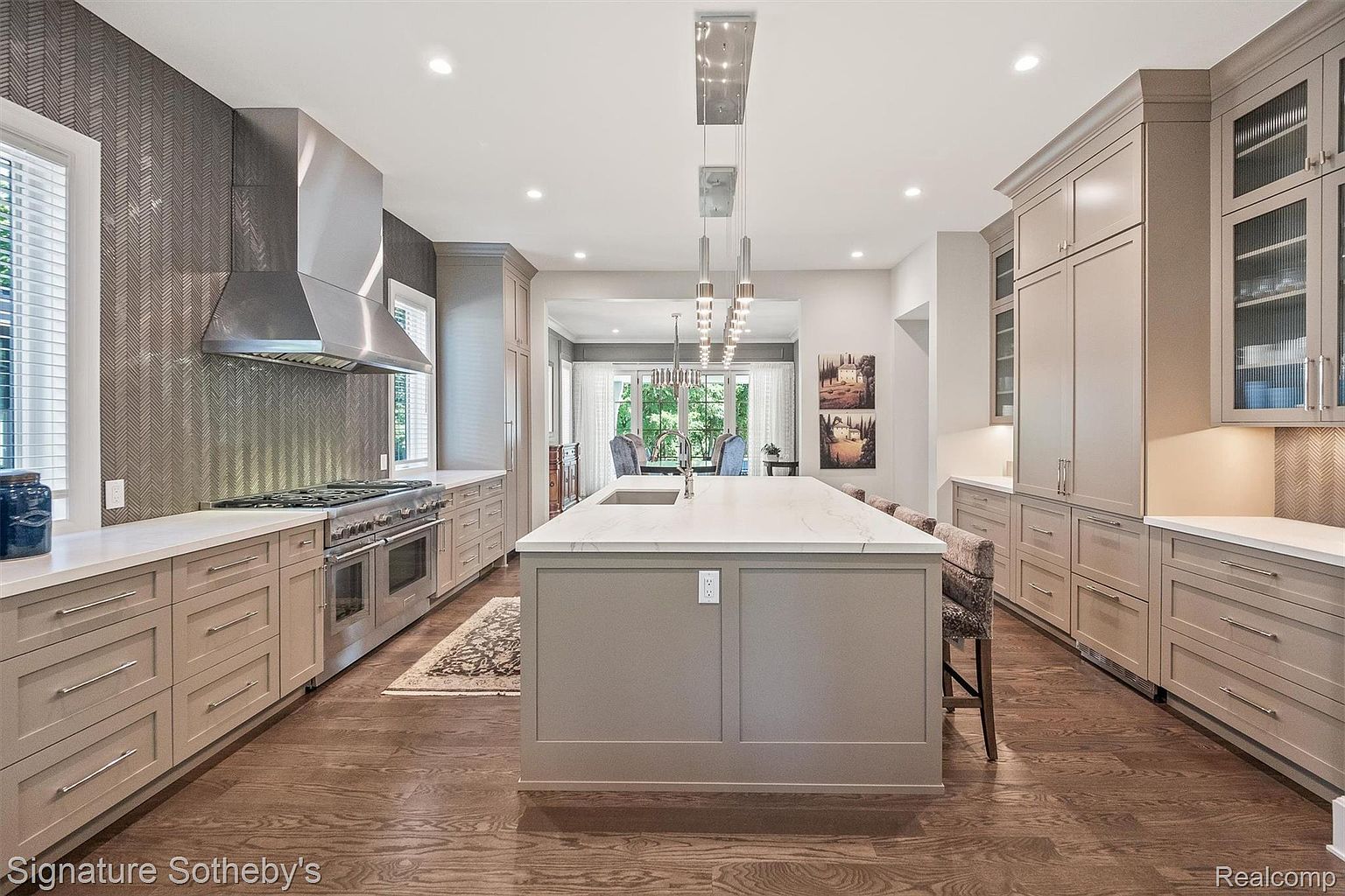 This is a bright and spacious kitchen featuring custom cabinetry in a neutral tone, a large center island with a white countertop, and stainless steel appliances. The kitchen is well-lit with recessed lighting and pendant lights above the island. The dark hardwood floors add warmth and contrast to the space, creating a luxurious and inviting atmosphere.