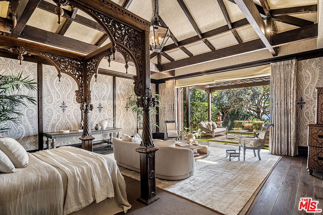 This luxurious primary bedroom features an ornate, dark wood carved canopy bed frame that serves as a dramatic focal point. The room seamlessly transitions to an expansive outdoor patio through large, open glass doors, blending indoor comfort with natural beauty. With its vaulted beamed ceiling, intricate paisley wallpaper, and sophisticated neutral furnishings, the space exudes an elegant, resort-like atmosphere.