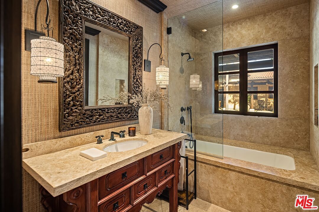 This elegant bathroom features a rich, dark wood vanity topped with a textured stone countertop and an ornate, carved-wood framed mirror. A glass-enclosed shower and tub combination is finished in matching stone tile, creating a cohesive and luxurious aesthetic. The warm lighting from the decorative wall sconces enhances the sophisticated, rustic-inspired atmosphere of the space.