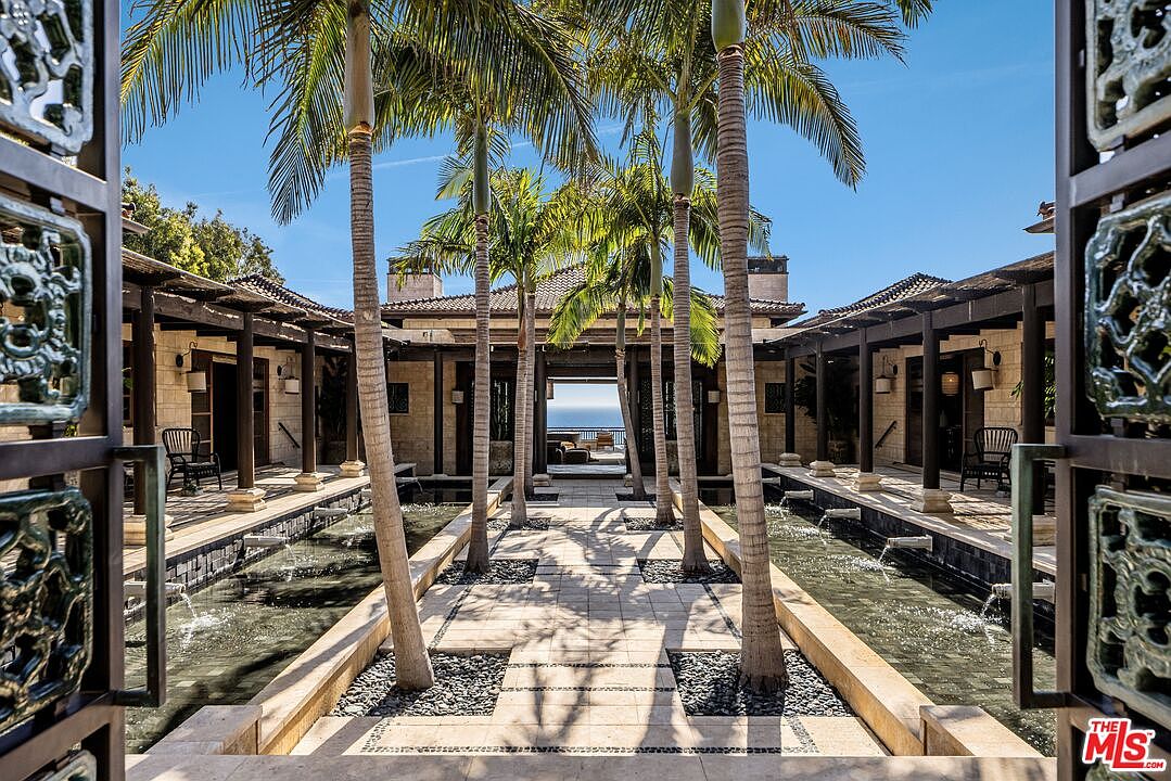 This stunning entryway features a symmetrical courtyard design with a central stone walkway flanked by tranquil reflecting pools and tall, elegant palm trees. The architectural style is sophisticated and resort-like, framed by ornate metal gates that open to reveal a clear view of the ocean in the distance. The perspective is centered and inviting, emphasizing the seamless transition between the luxurious home and the natural coastal environment.