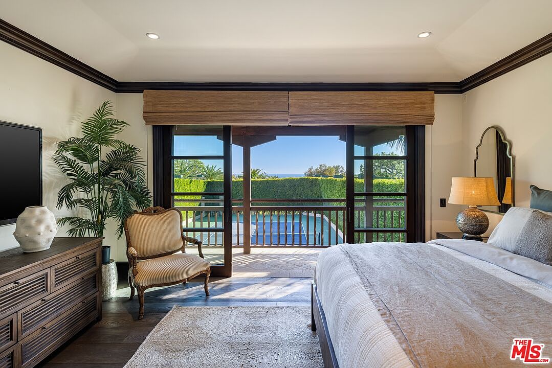 This elegant primary bedroom features a neutral color palette, dark wood flooring, and a large sliding glass door that opens onto a private balcony overlooking a tennis court and lush greenery. The room is furnished with a comfortable bed, a classic striped armchair, and a wooden dresser, creating a serene and sophisticated atmosphere. The high tray ceiling and warm lighting enhance the inviting, upscale feel of the space.