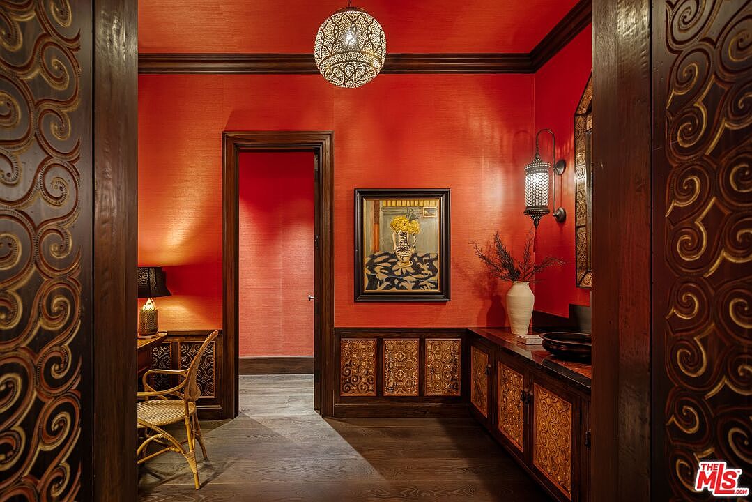 This opulent hallway features striking red textured wallpaper, dark wood trim, and intricate carved wooden panels that frame the space. A decorative metal pendant light hangs from the ceiling, casting a warm glow over the dark hardwood floors and a framed floral painting. The perspective is centered through a doorway, highlighting the rich, exotic aesthetic and sophisticated craftsmanship of the home's interior design.