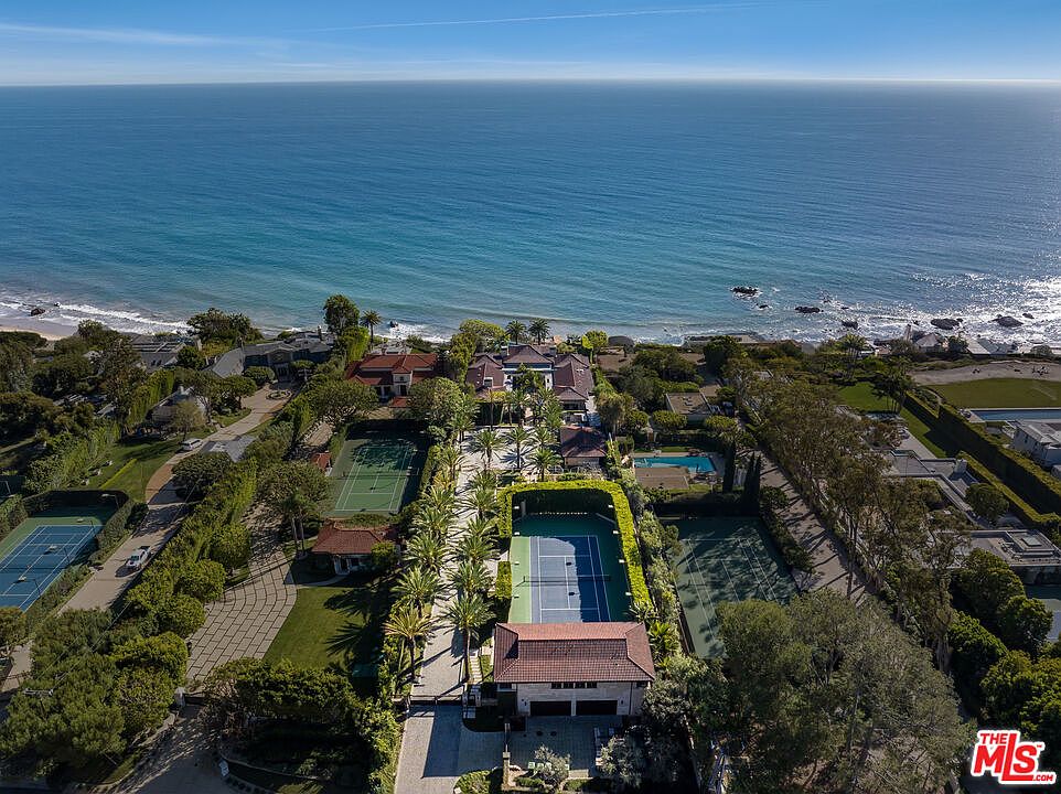This stunning aerial view captures a sprawling, luxurious coastal estate situated directly on the oceanfront. The property features multiple tennis courts, lush manicured landscaping, and a grand main residence, all set against the backdrop of the vast, blue Pacific Ocean. The perspective highlights the expansive scale of the grounds and the prime, secluded location of this high-end real estate.