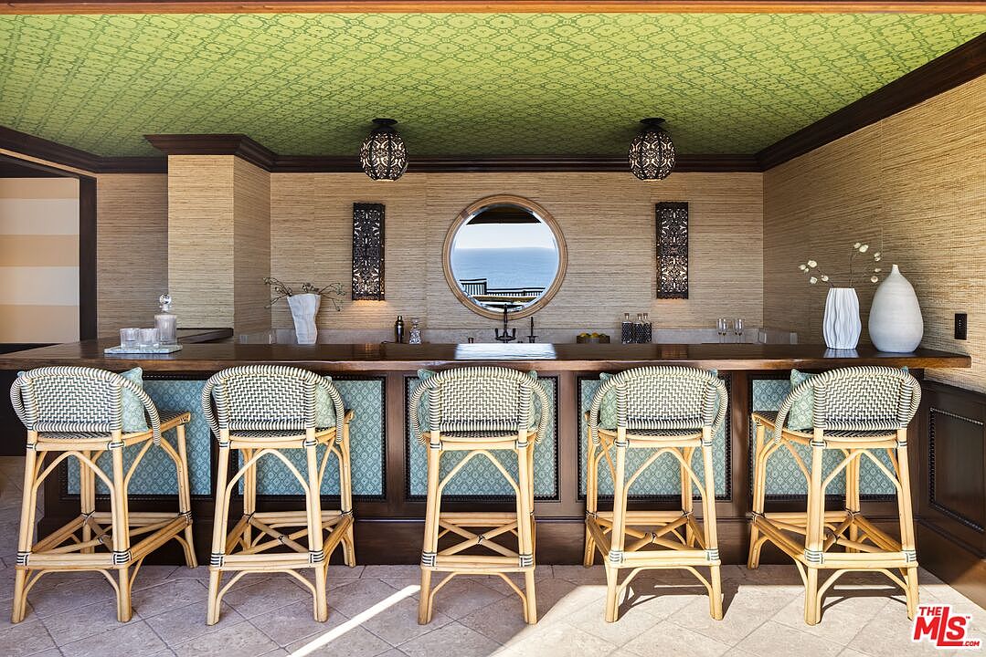 This sophisticated home bar area features a long wooden counter with five woven rattan bar stools, set against a textured grasscloth wall. A circular window provides a scenic view of the ocean, while intricate patterned wallpaper on the ceiling and decorative wall sconces add a touch of global-inspired elegance. The space is designed for entertaining, blending natural textures with a refined, coastal aesthetic.