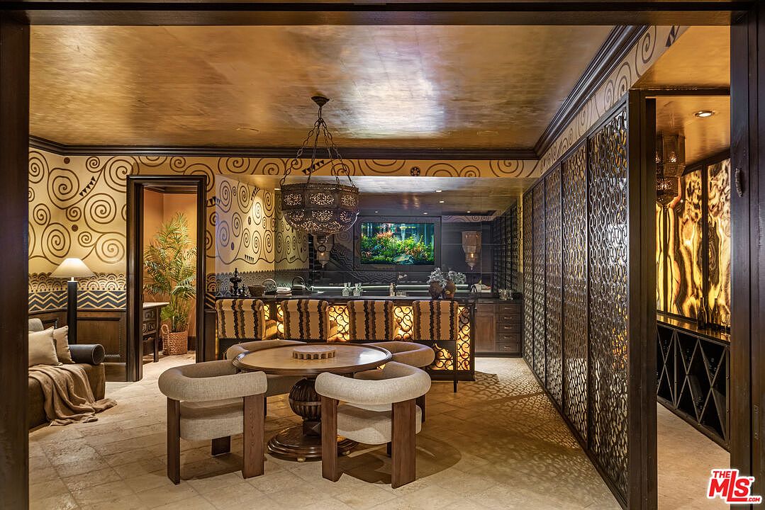This luxurious, exotic-themed basement lounge features a central circular game table surrounded by four upholstered chairs, set against a backdrop of intricate, patterned wallpaper and a metallic-finished ceiling. A fully equipped bar with illuminated stools and a decorative metal screen partition adds to the sophisticated, moody atmosphere. The space is designed for high-end entertainment, blending ornate textures with warm, ambient lighting to create an exclusive, club-like feel.