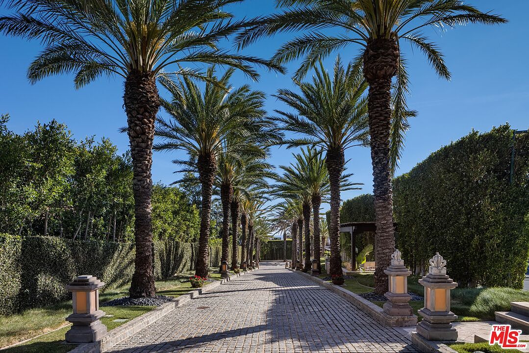 This grand, gated driveway features a long, cobblestone path lined with majestic, mature palm trees that create a dramatic and symmetrical entrance. The property is enclosed by tall, manicured hedges, providing a sense of privacy and exclusivity, while decorative stone lanterns illuminate the walkway. The perspective is a low-angle, eye-level shot looking down the center of the path, emphasizing the scale and luxurious atmosphere of the estate.
