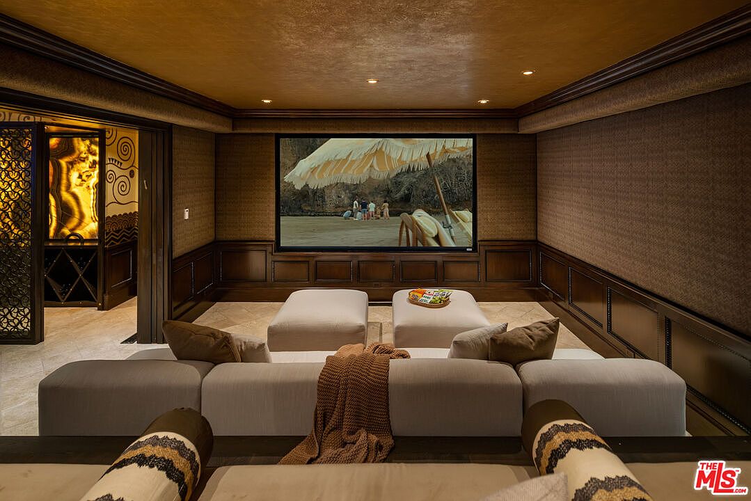 This luxurious home theater features a large projection screen, plush tiered seating with a neutral-toned sectional, and elegant wood-paneled walls. The room is accented by a warm, ambient ceiling finish and leads into a sophisticated, illuminated wine cellar area visible through the doorway. The overall aesthetic is one of refined comfort and high-end entertainment, perfect for a cinematic viewing experience.