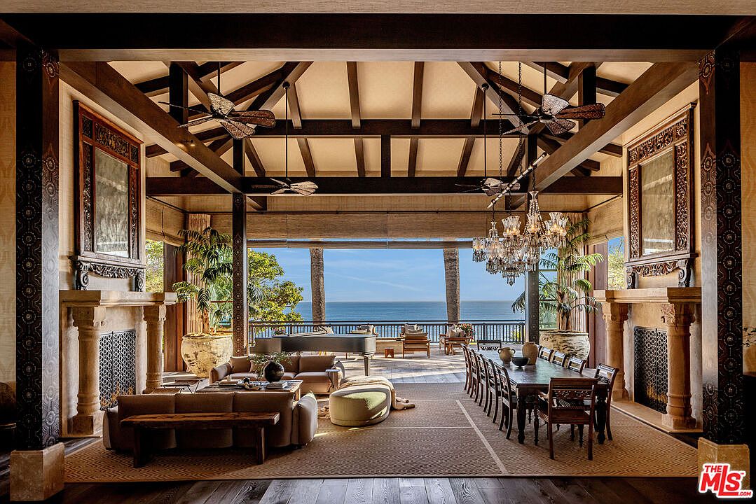 This expansive, open-concept great room seamlessly blends a sophisticated living area with a formal dining space, all under a dramatic vaulted ceiling with exposed timber beams. The room features dual stone fireplaces, ornate carved wood accents, and a grand piano, all oriented toward a massive, unobstructed view of the ocean through a wide-open terrace. The aesthetic is a luxurious, tropical-inspired coastal retreat, characterized by rich textures, warm wood tones, and an airy, indoor-outdoor flow.
