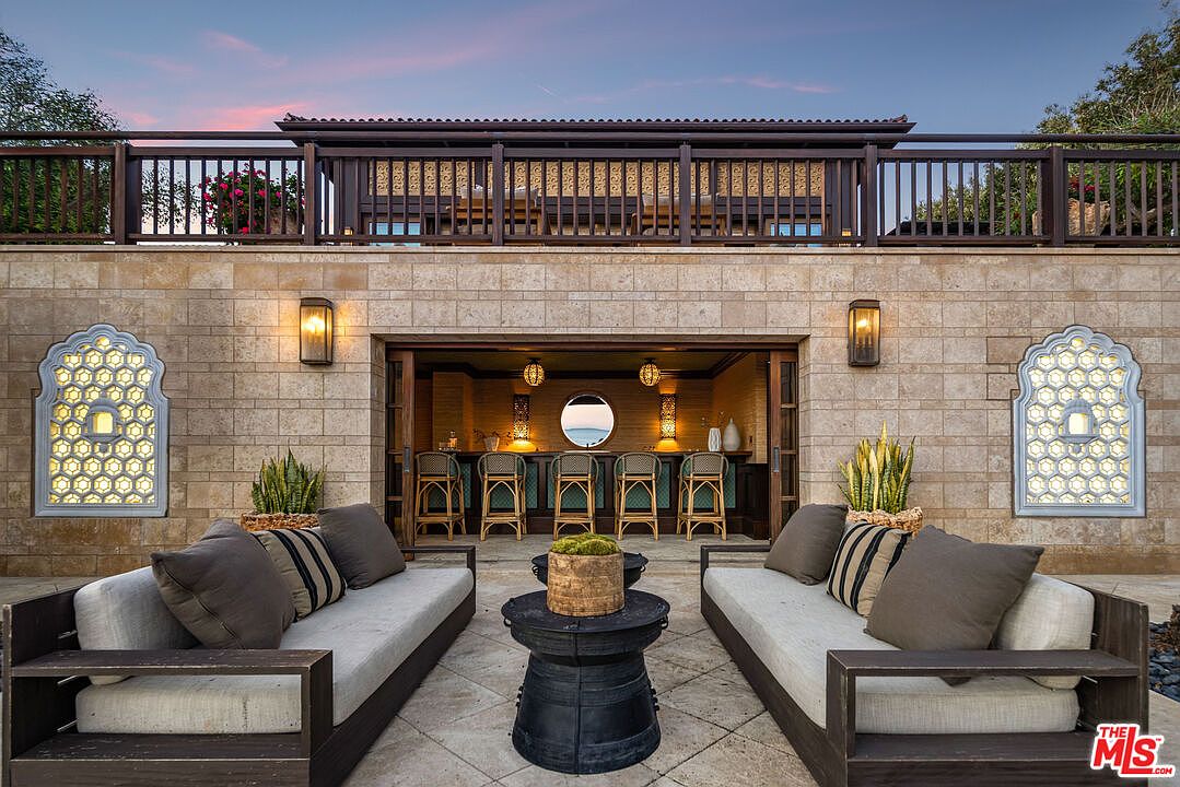 This elegant outdoor patio space features a symmetrical layout with two plush, modern sofas facing a central drum-style coffee table. The area is anchored by a stone-walled bar alcove with barstool seating, illuminated by warm wall sconces and decorative lattice windows. The perspective is a centered, eye-level shot that highlights the sophisticated, resort-like atmosphere and the seamless transition between the lounge area and the bar.