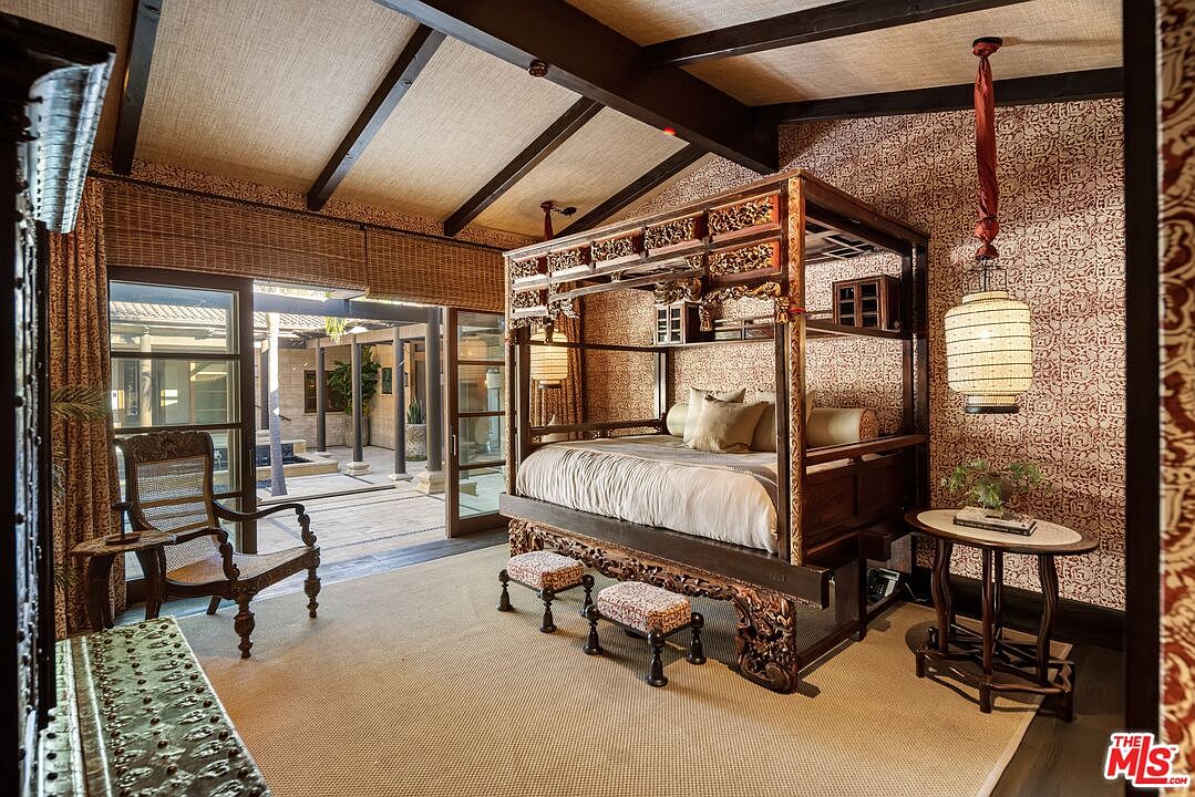 This primary bedroom features an ornate, dark wood canopy bed with intricate carvings, serving as the room's dramatic focal point. The space is decorated with patterned wallpaper, exposed ceiling beams, and a neutral-toned area rug, while large glass doors open directly onto a serene, Asian-inspired courtyard. The overall aesthetic is one of luxurious, exotic elegance, blending traditional craftsmanship with a seamless indoor-outdoor living experience.