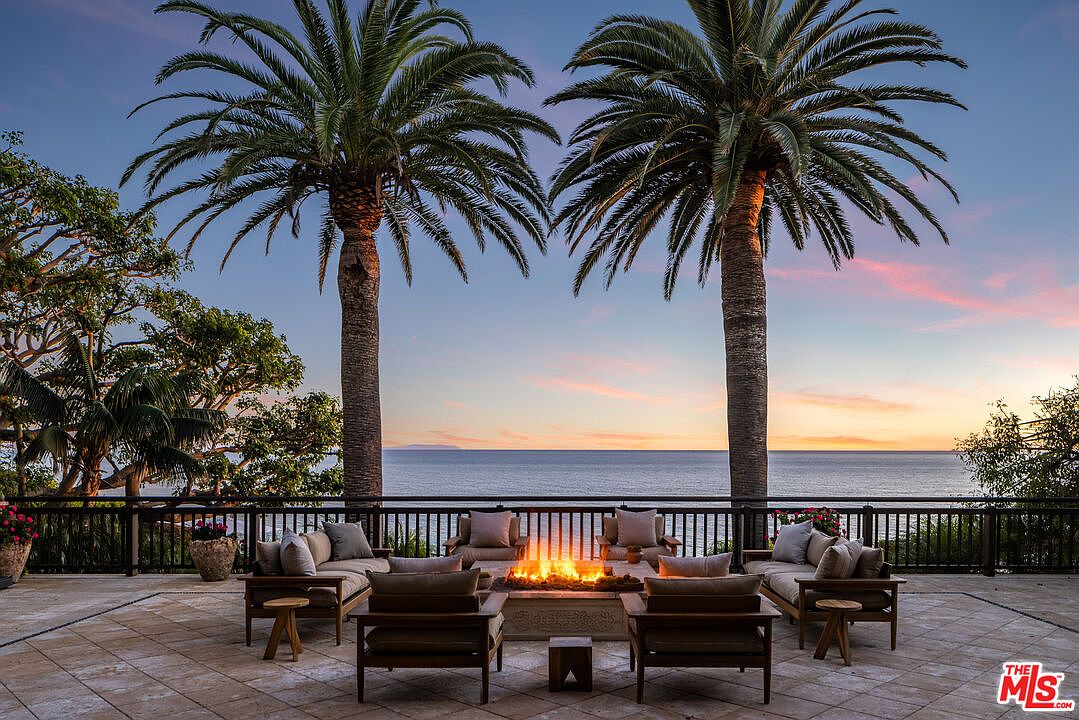This stunning outdoor patio offers a luxurious lounge area centered around a glowing fire pit, perfectly positioned to capture panoramic ocean views at sunset. The space is framed by two majestic palm trees and features comfortable, high-end seating arrangements on a stone-tiled terrace. The serene atmosphere and unobstructed horizon create an elegant, resort-like setting ideal for evening relaxation.