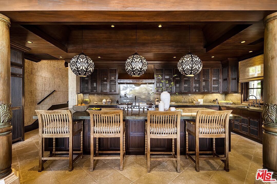 This luxurious, tropical-inspired kitchen features rich, dark wood cabinetry and a large central island topped with a stone countertop, complemented by four wooden bar stools. Three ornate, spherical pendant lights hang from a coffered wood-paneled ceiling, casting a warm glow over the space. The room is framed by decorative columns and stone flooring, creating an elegant and sophisticated atmosphere.