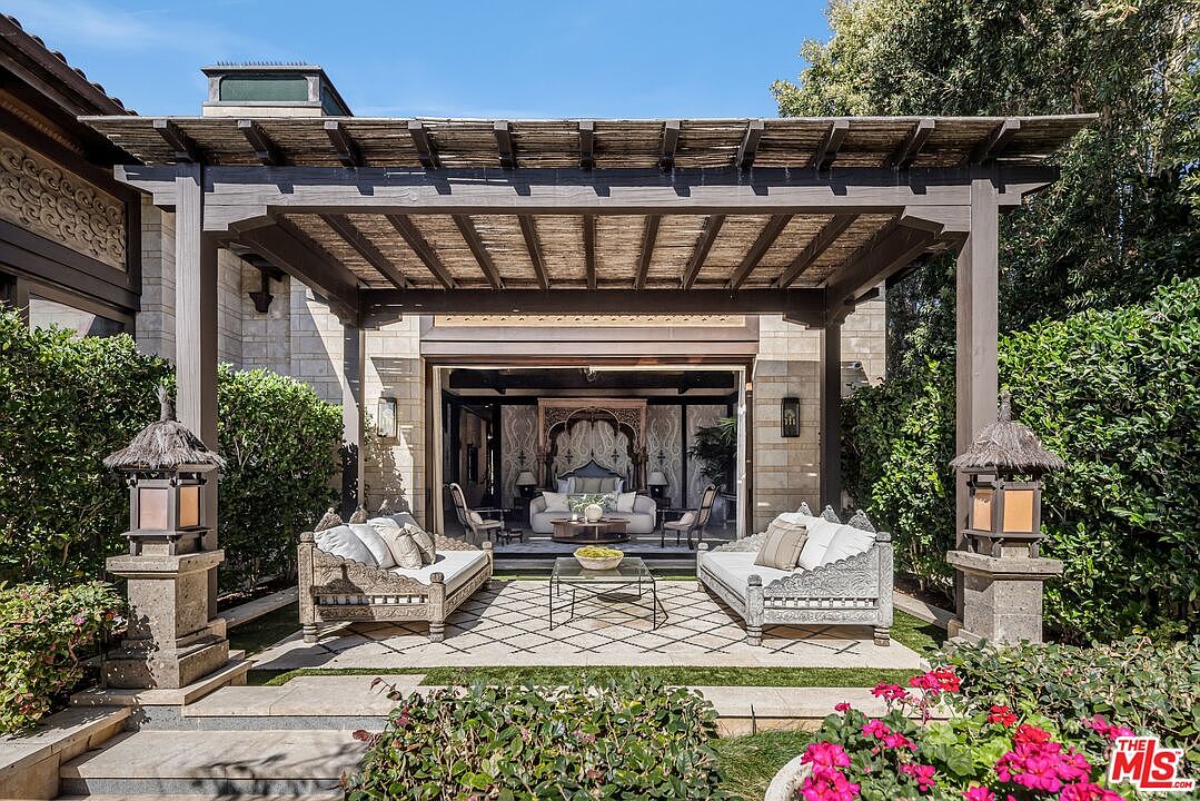 This luxurious outdoor patio features a large, dark-stained wooden pergola with a woven reed roof, providing shade over a sophisticated seating area. Two ornate, carved wooden sofas with plush white cushions face each other around a glass-topped coffee table, all set upon a patterned stone patio. The space seamlessly transitions into an indoor living area through wide-open glass doors, showcasing an elegant, exotic-inspired interior design.
