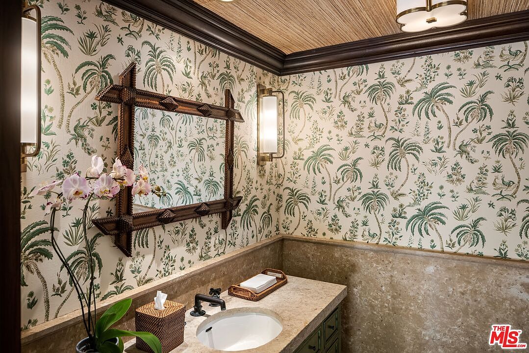This elegant powder room features sophisticated palm-leaf patterned wallpaper and a textured grasscloth ceiling, creating a refined tropical aesthetic. The space is anchored by a custom stone vanity with a dark, ornate wooden mirror and matching wall-mounted light fixtures. The combination of natural stone textures and classic botanical prints gives the room a luxurious, boutique-hotel feel.