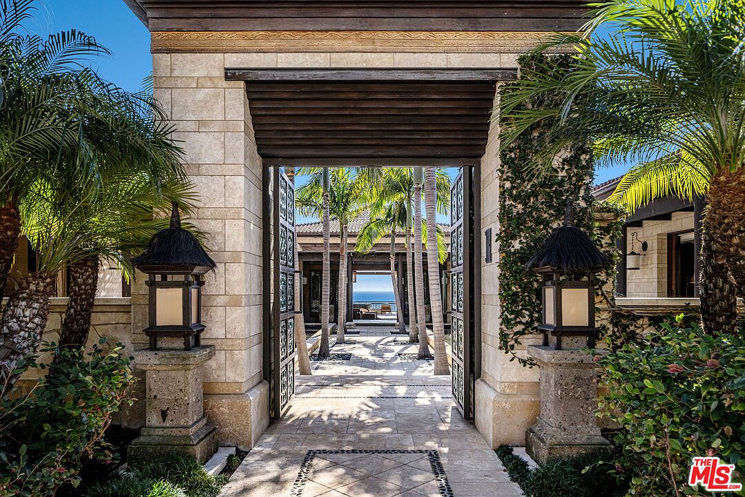 This grand, resort-style entryway features a massive stone-clad gatehouse with intricate wooden detailing and two ornate, lantern-style light fixtures flanking the entrance. The open iron gates reveal a serene, palm-lined courtyard path that leads directly to a stunning, unobstructed view of the ocean horizon. The architectural style blends tropical luxury with Mediterranean influences, creating an inviting and sophisticated first impression.