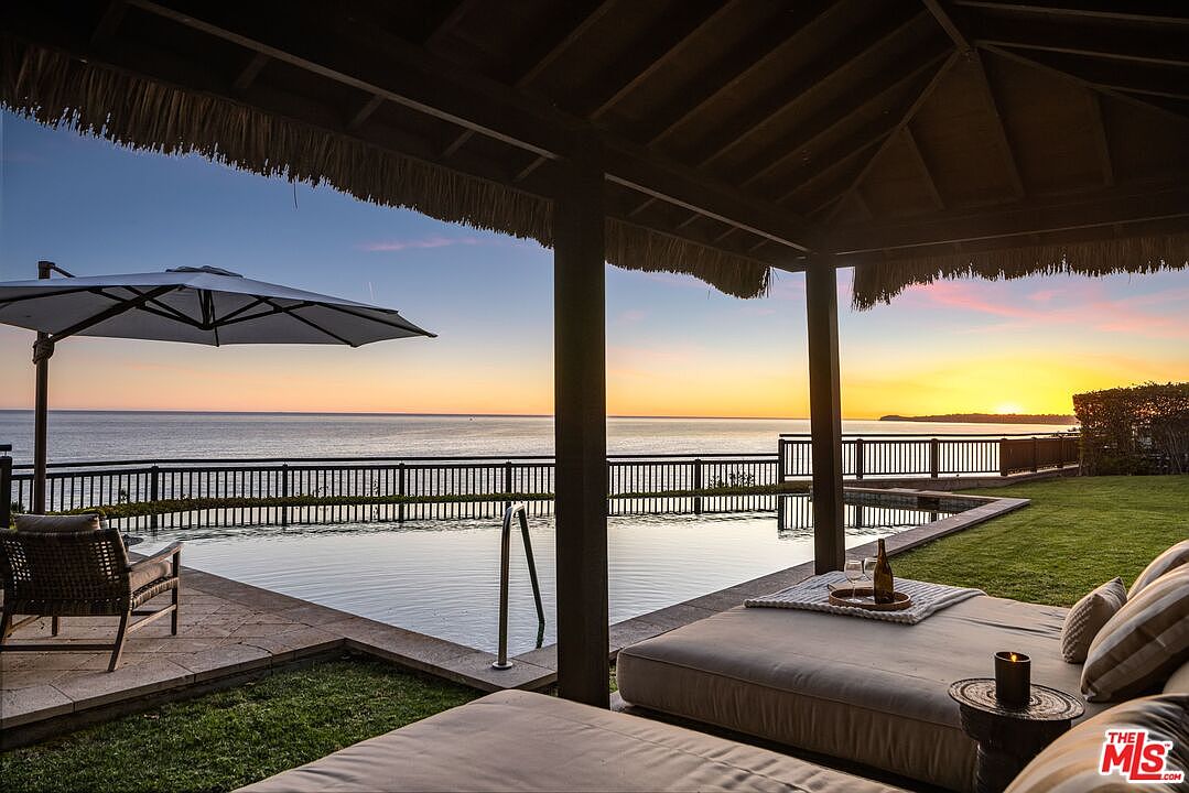 This stunning outdoor living space features a luxurious infinity-edge pool overlooking a breathtaking sunset over the ocean. The covered patio area includes comfortable lounge seating and a thatched-roof structure, creating a serene and high-end resort-like atmosphere. The perspective captures the seamless transition between the private pool deck and the expansive coastal horizon.