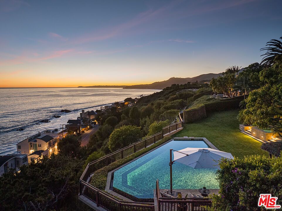 This stunning aerial perspective captures a luxurious infinity-edge pool overlooking a dramatic coastal sunset in Malibu. The scene features a lush green lawn, a modern patio area with a built-in fire feature, and a panoramic view of the Pacific Ocean meeting the rugged coastline. The composition emphasizes the seamless integration of high-end outdoor living with the natural beauty of the California shore.
