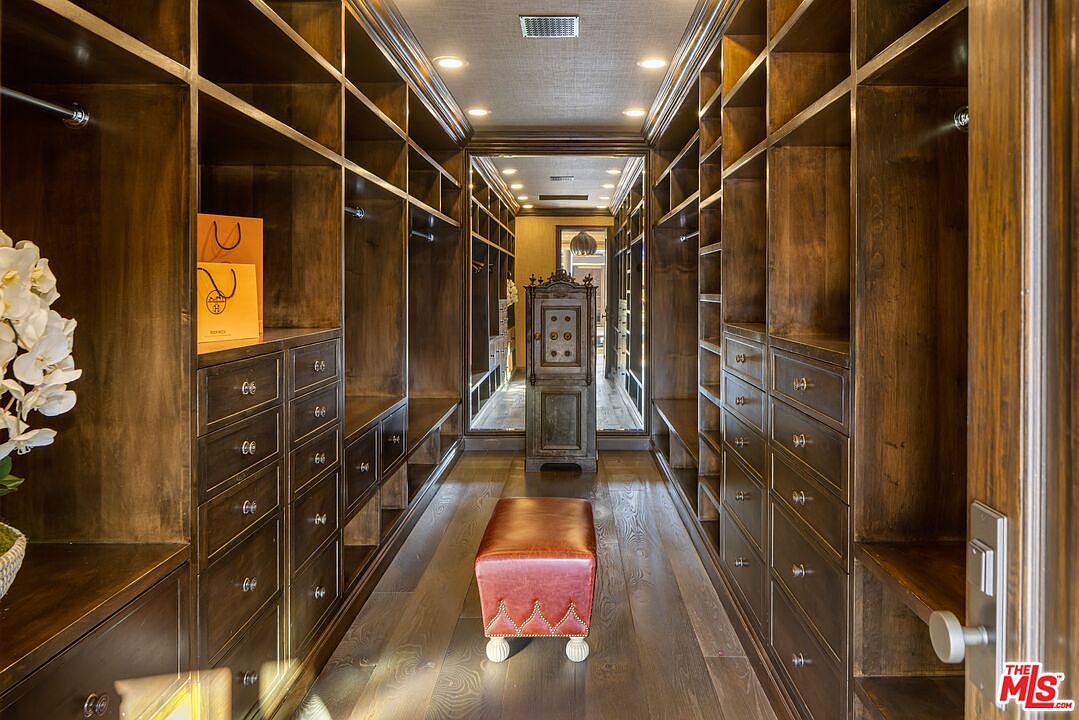 This luxurious walk-in closet features rich, dark-stained custom cabinetry with extensive shelving and drawer storage lining both walls. A central red leather ottoman provides a sophisticated focal point, while a large mirror at the end of the hallway creates an expansive, symmetrical perspective. The warm recessed lighting and high-end finishes contribute to an elegant and organized atmosphere.
