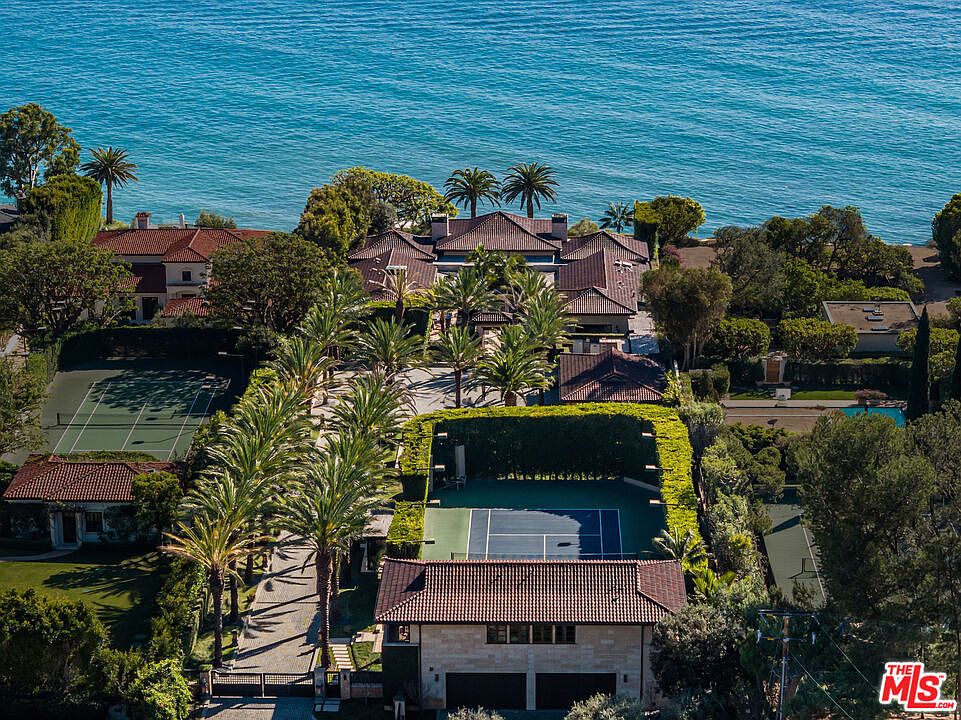 This high-angle aerial view captures a sprawling, luxurious coastal estate featuring a primary residence with a classic tile roof, multiple tennis courts, and lush, manicured landscaping. The property is situated directly along the oceanfront, showcasing a seamless blend of private recreational amenities and expansive outdoor living spaces. The perspective emphasizes the grand scale of the estate and its prime, secluded location overlooking the water.
