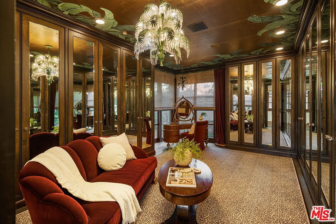 This luxurious walk-in closet features dark wood cabinetry with mirrored doors, creating a sophisticated and expansive atmosphere. The room is accented by a vibrant red velvet sofa, a central oval coffee table, and a dramatic palm-leaf chandelier hanging from a ceiling painted with tropical foliage. The space is further enhanced by a vanity area and a patterned carpet, blending high-end fashion storage with a comfortable lounge aesthetic.