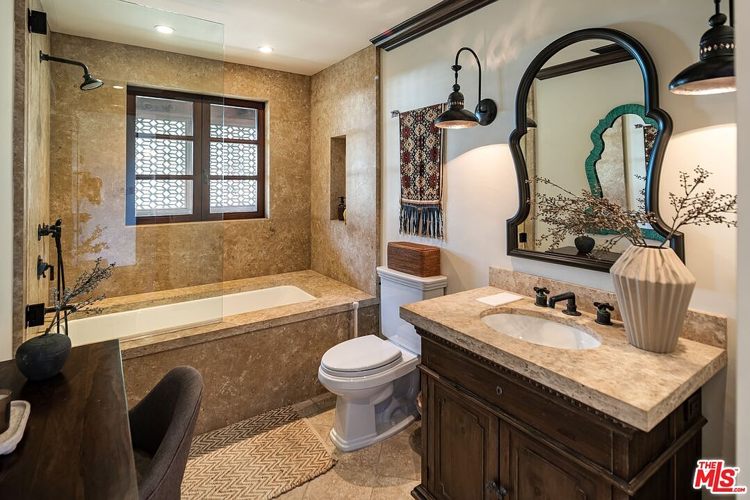 This elegant bathroom features a luxurious stone-tiled bathtub and shower combination, complemented by a rustic wooden vanity with a stone countertop. The space is accented with a decorative wall hanging, a unique arched mirror, and warm lighting, creating a sophisticated and inviting atmosphere. The perspective captures the entire room, highlighting the blend of natural textures and refined design elements.